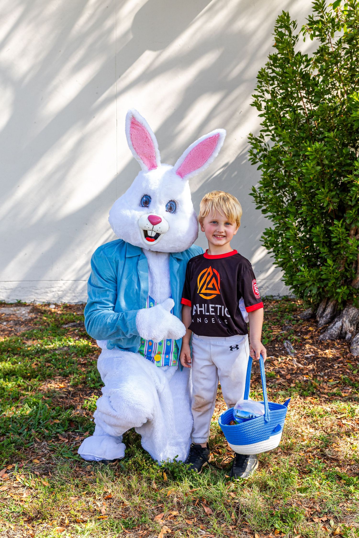 Young boy holding blue Easter basket poses with Easter Bunny mascot in blue jacket outdoors.