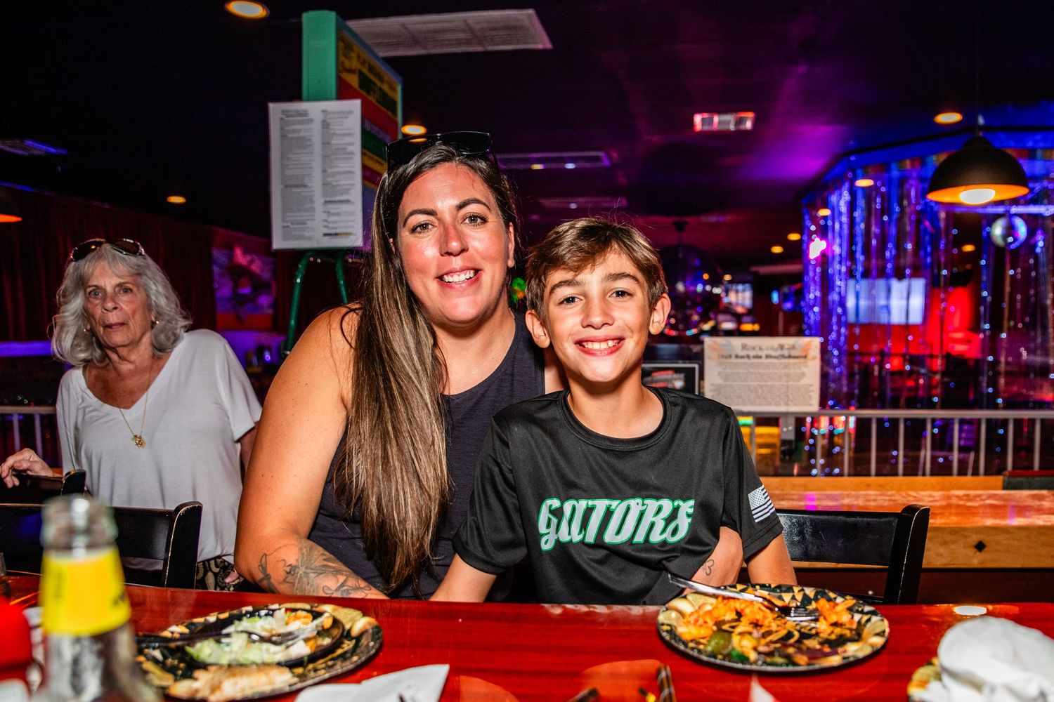 Woman and boy smiling at restaurant table with pizza and food, colorful bar lighting in background.