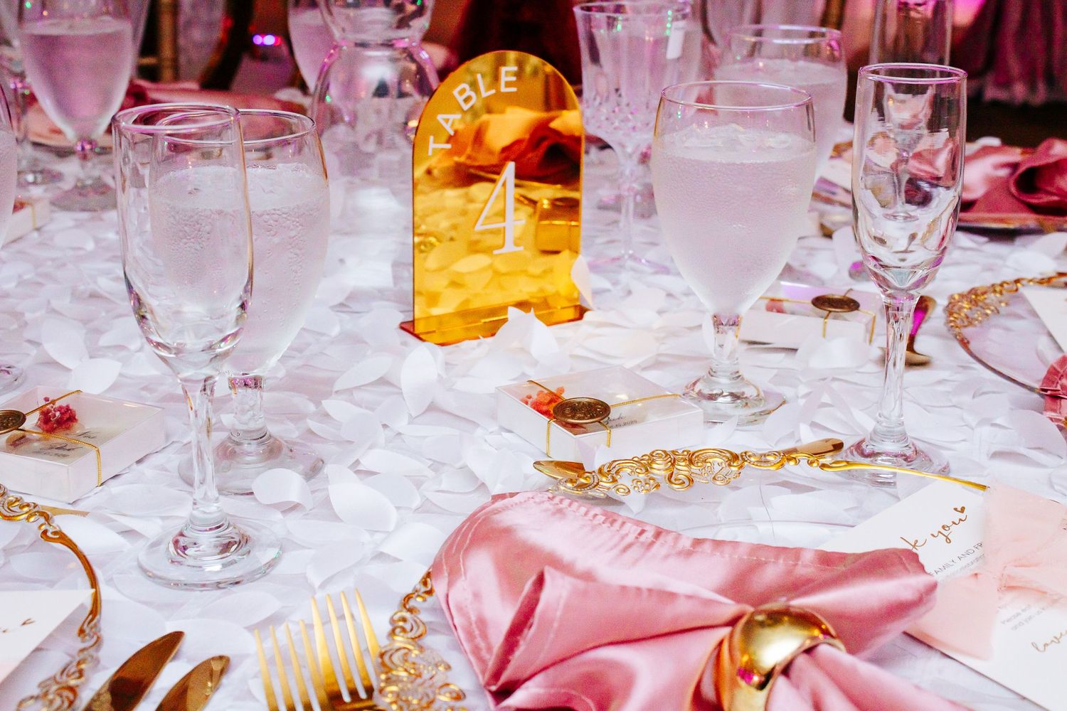 Pink and gold table setting with wine glasses and amber lighting at formal event.