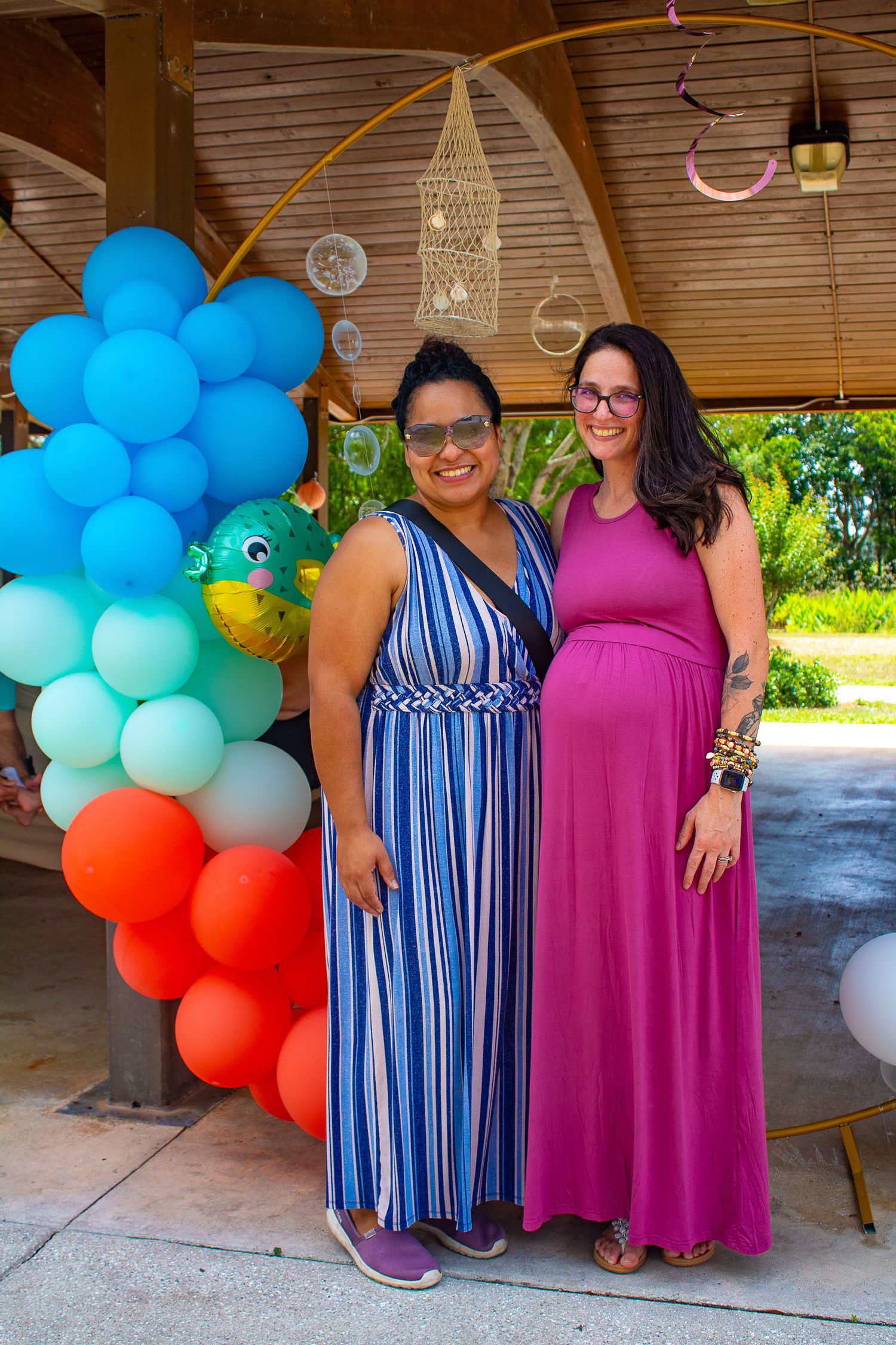 Baby shower celebration with balloon arch decor in blue, teal and orange colors under wooden pavilion.