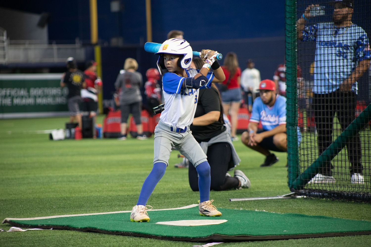 Baseball player practices batting stance on green training mat near batting cage.
