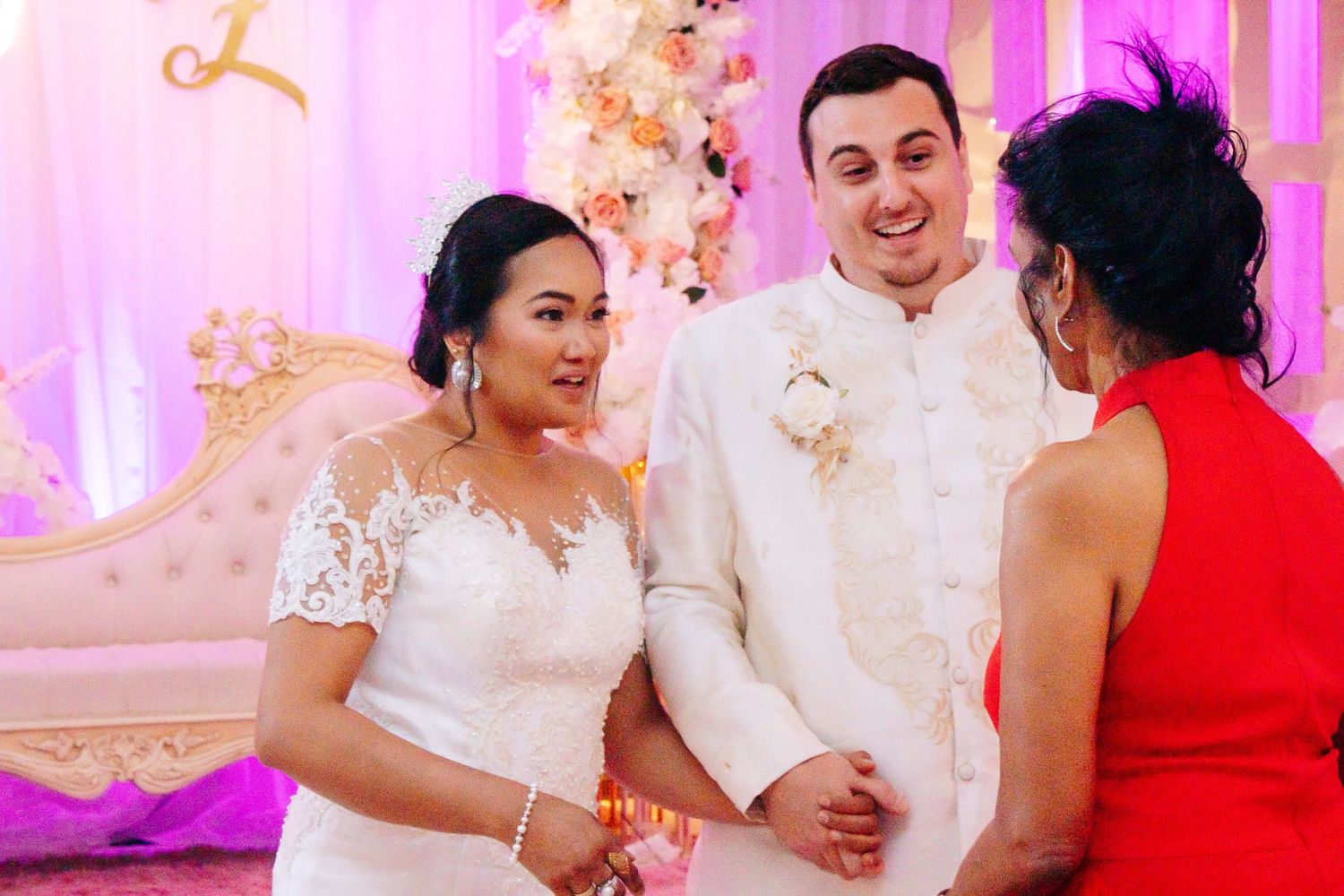Wedding couple shares a moment with guest in red dress against pink backdrop.