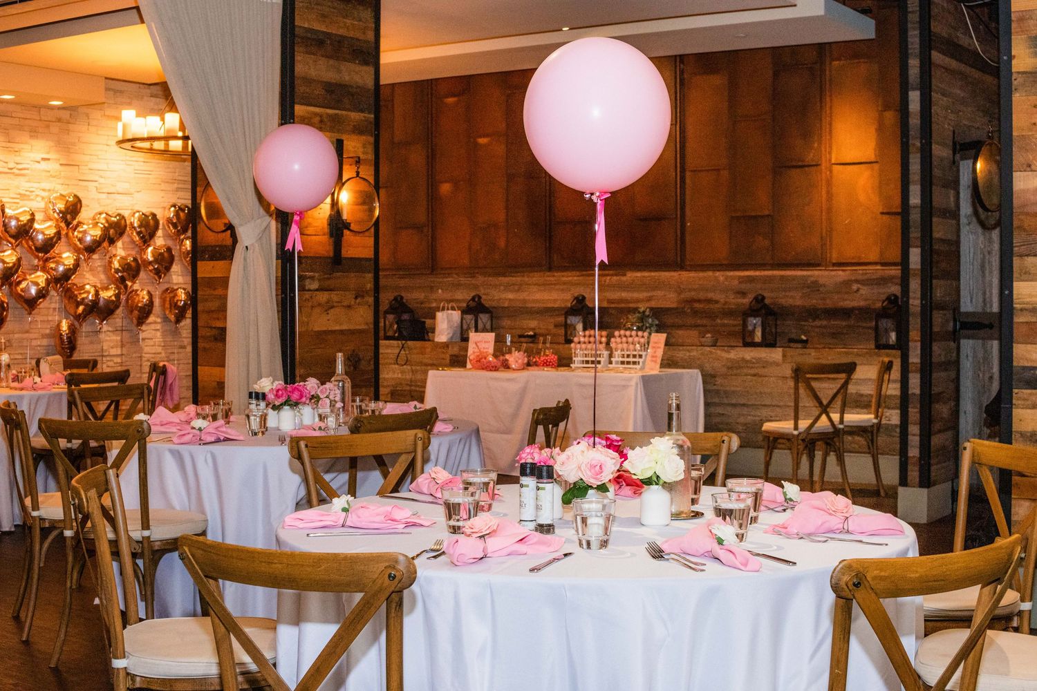 Round tables decorated with white linens, pink balloons, and cross-back chairs in a warm-lit venue.