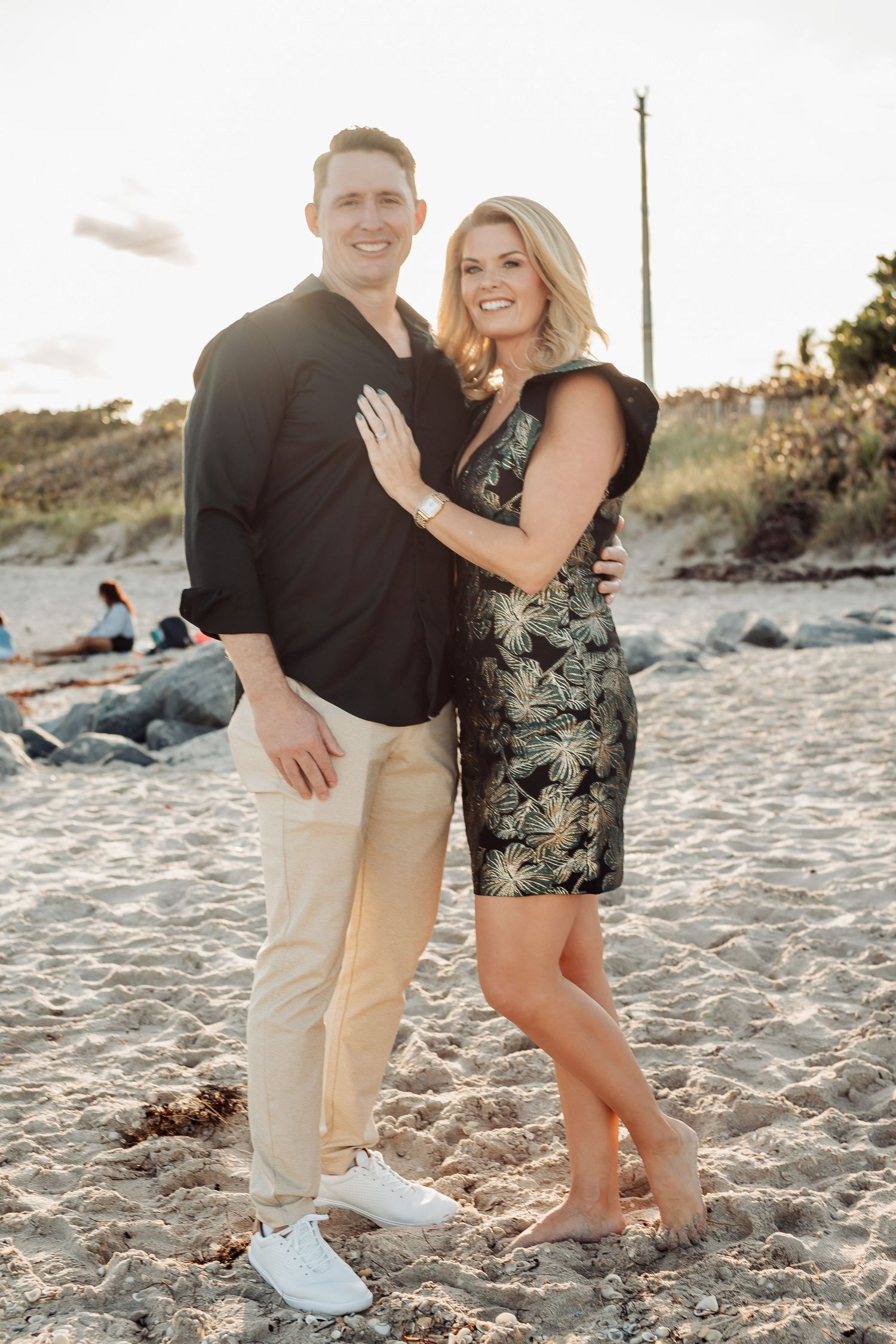 Couple in elegant attire embracing on sandy beach during golden hour photoshoot.