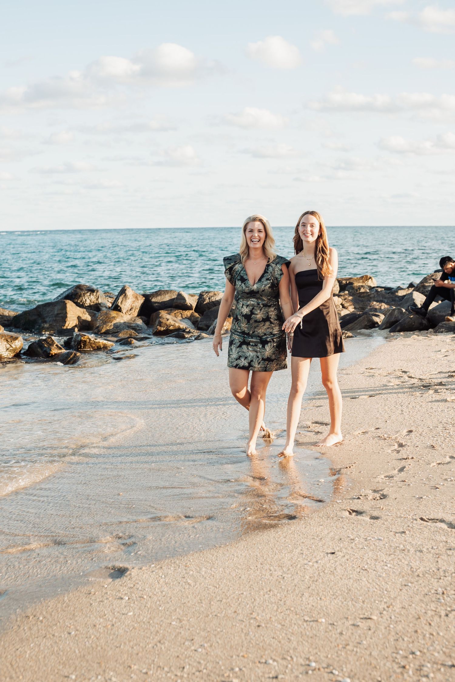 Two friends walking barefoot along beautiful beach with rocky shoreline.