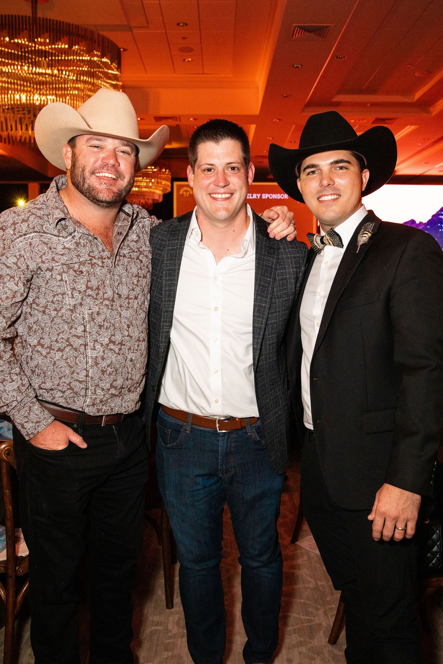 Three guests in cowboy hats and formal wear pose together at an evening function.