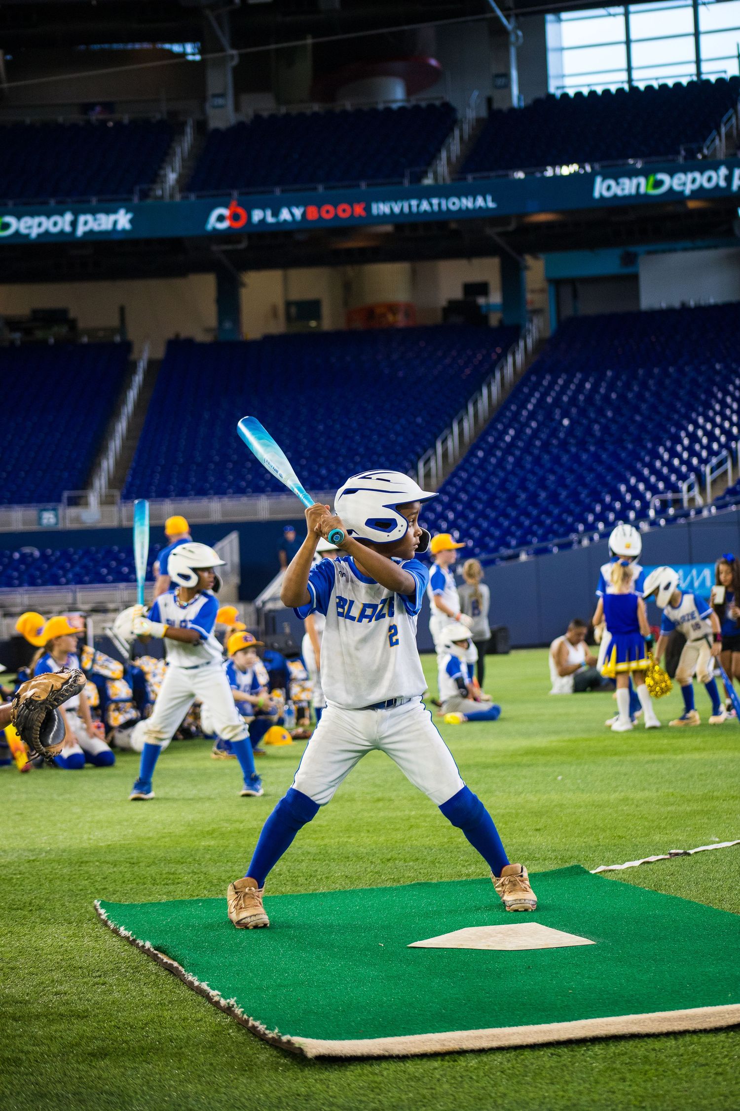Baseball mascot demonstrating batting stance on practice mat at empty stadium field.