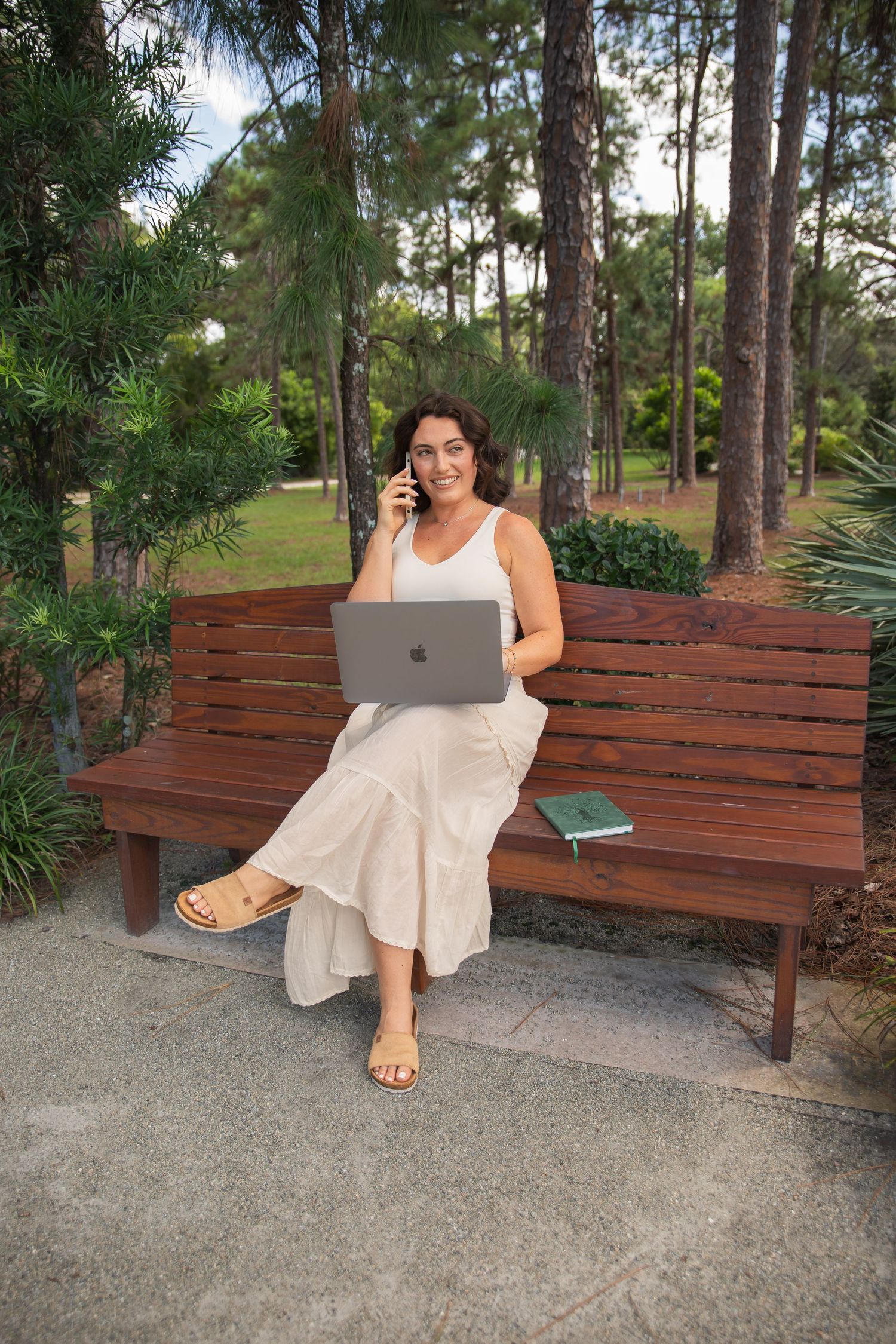 A woman in a white sundress sits on a wooden park bench beneath tall pine trees.