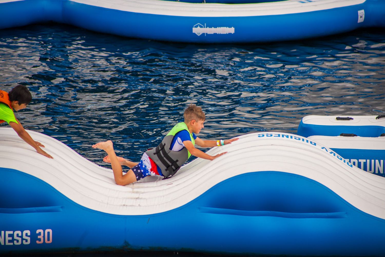 People enjoying an inflatable water obstacle course with blue floating platforms at an outdoor pool.