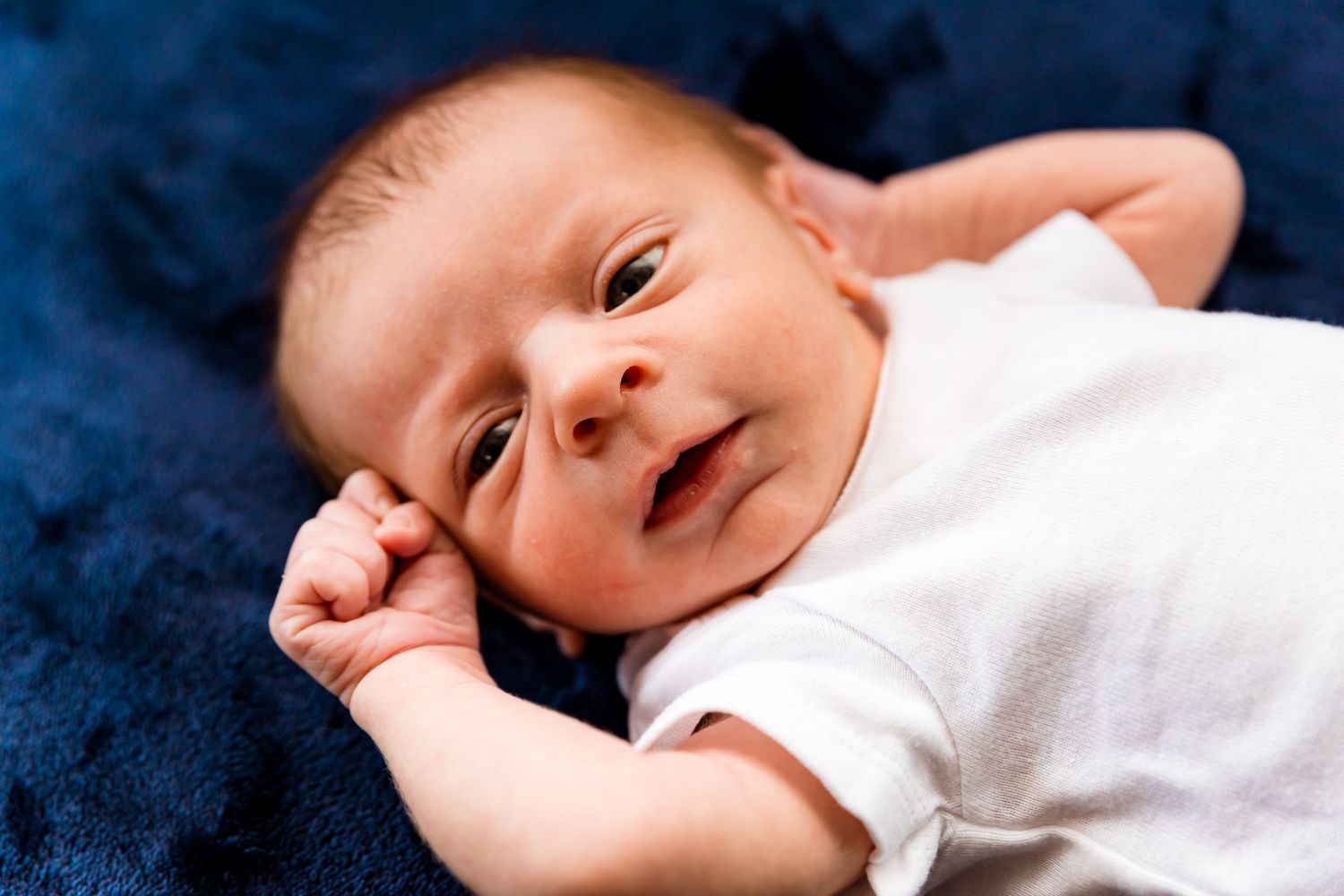 Newborn baby in white onesie laying on dark blue blanket looking up with curious expression.