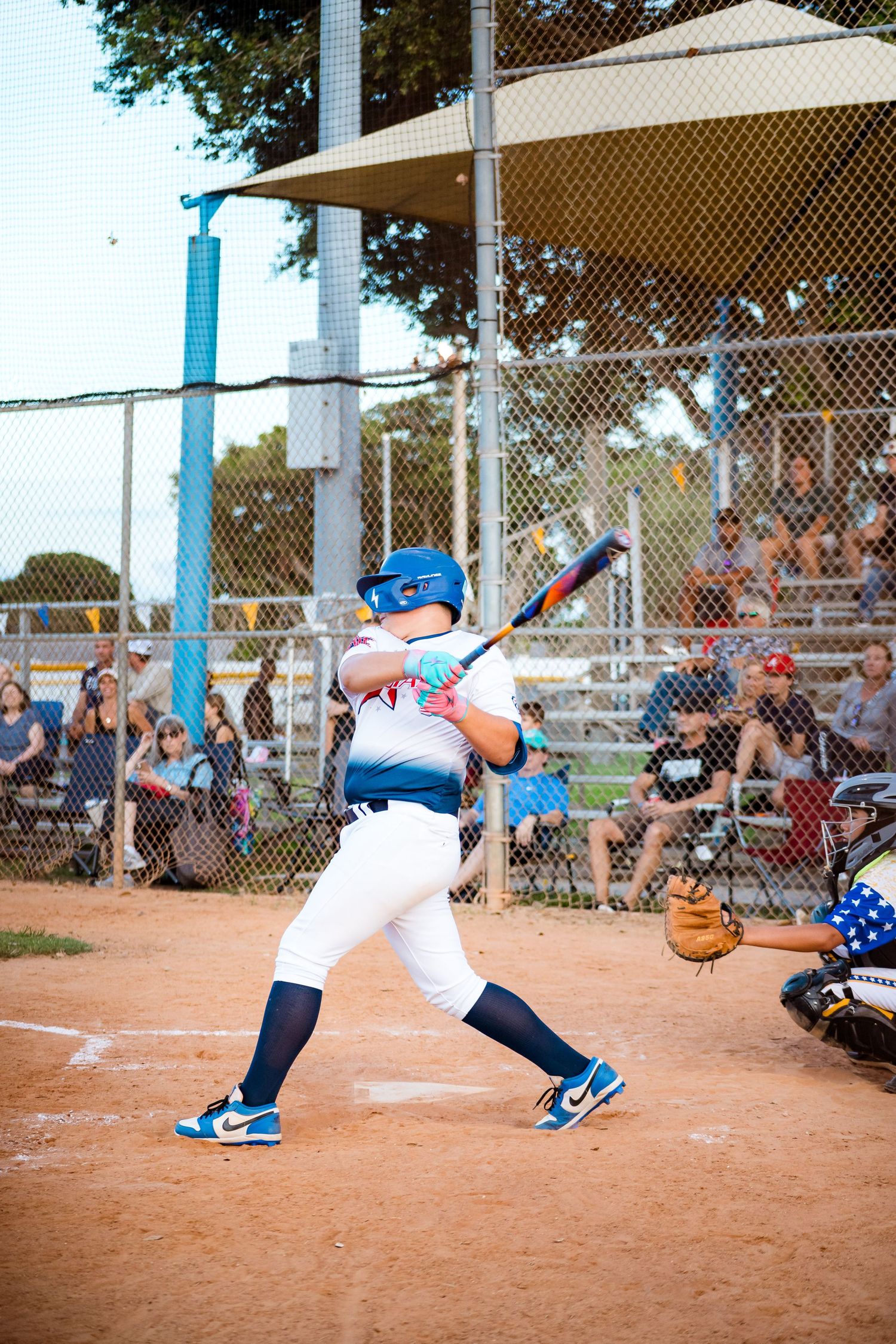 Youth baseball player in blue helmet takes batting stance at home plate during game with spectators in background.