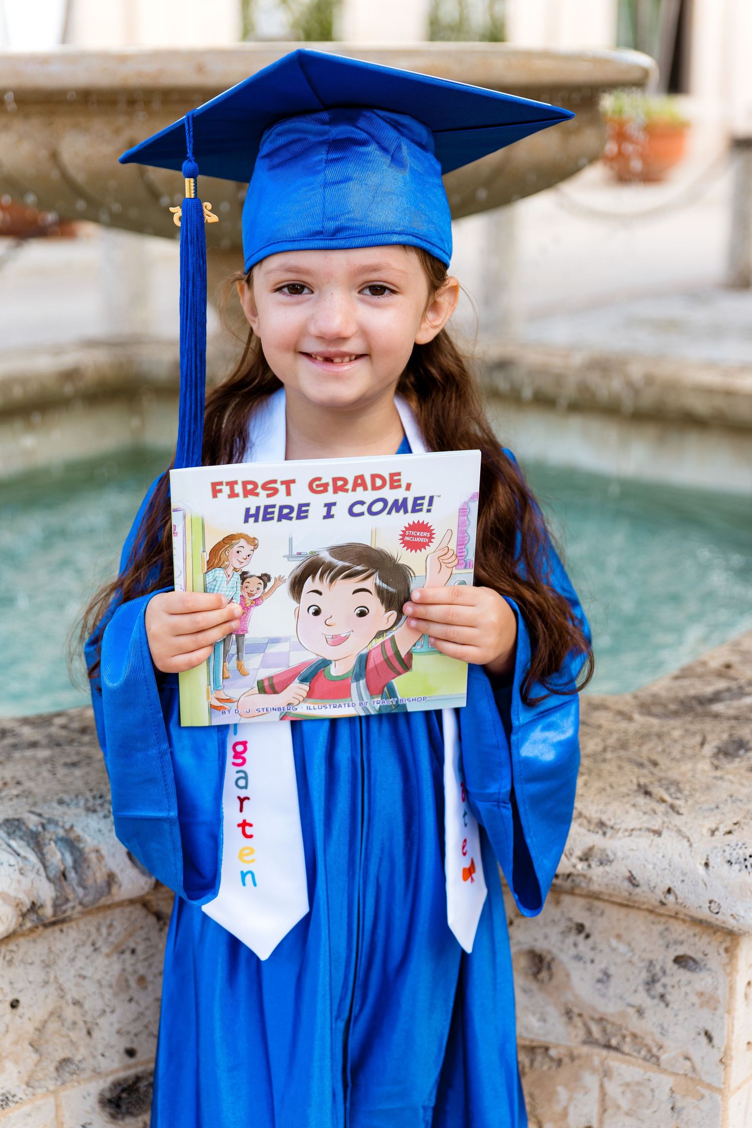 A small graduate in bright blue cap and gown holds up a children's book titled 'First Grade Here I Come' by a fountain.