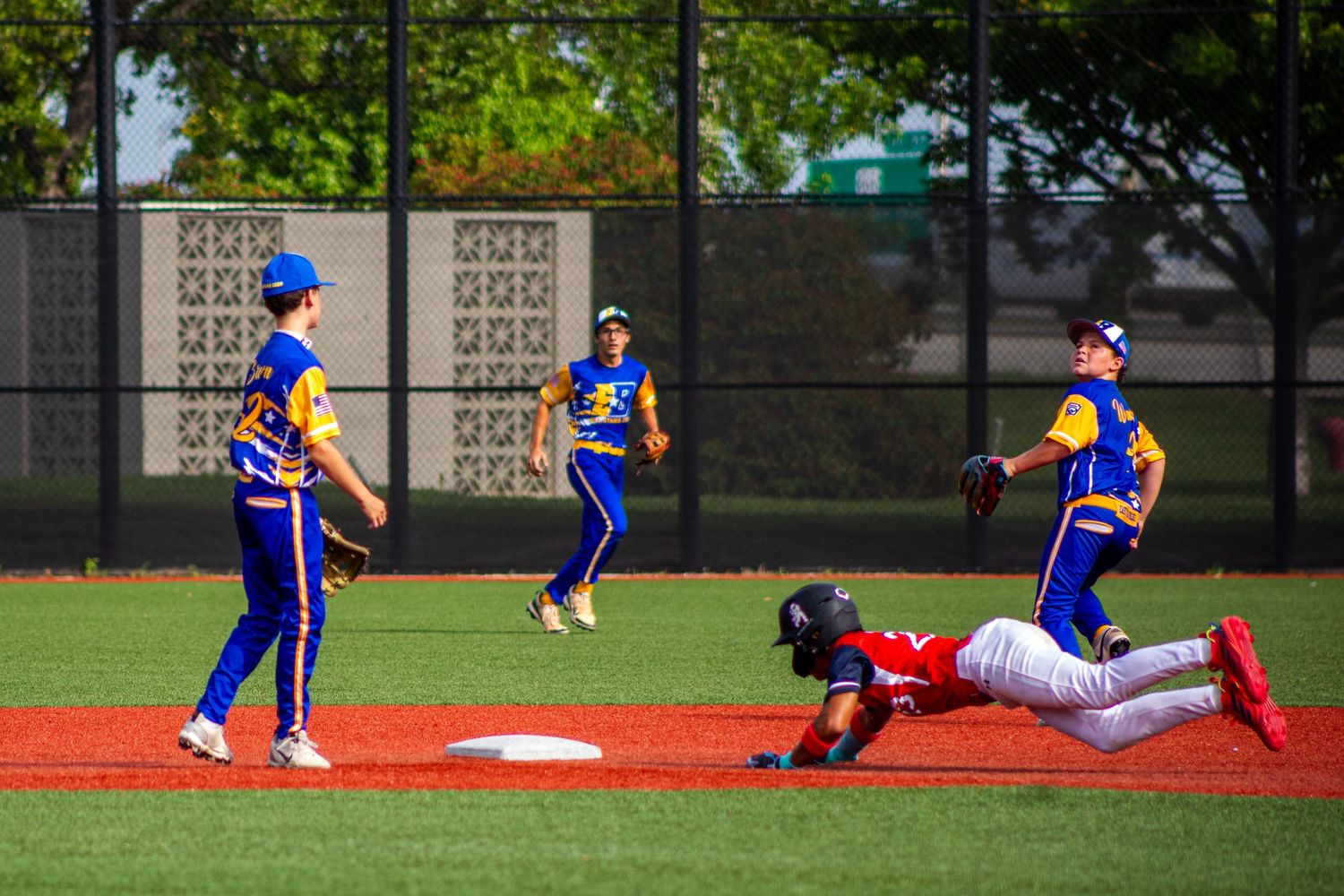 Baseball player sliding into base while three opponents in blue and yellow uniforms attempt a tag during a game.