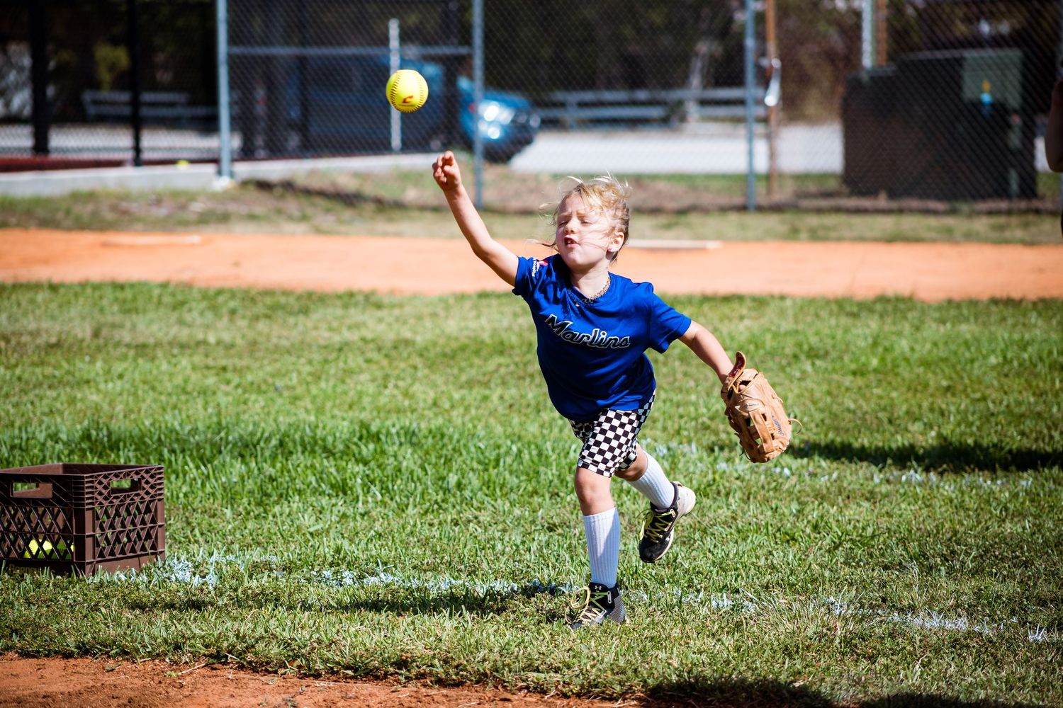 Someone in a blue shirt and baseball glove throws softball during outdoor practice on grassy field.