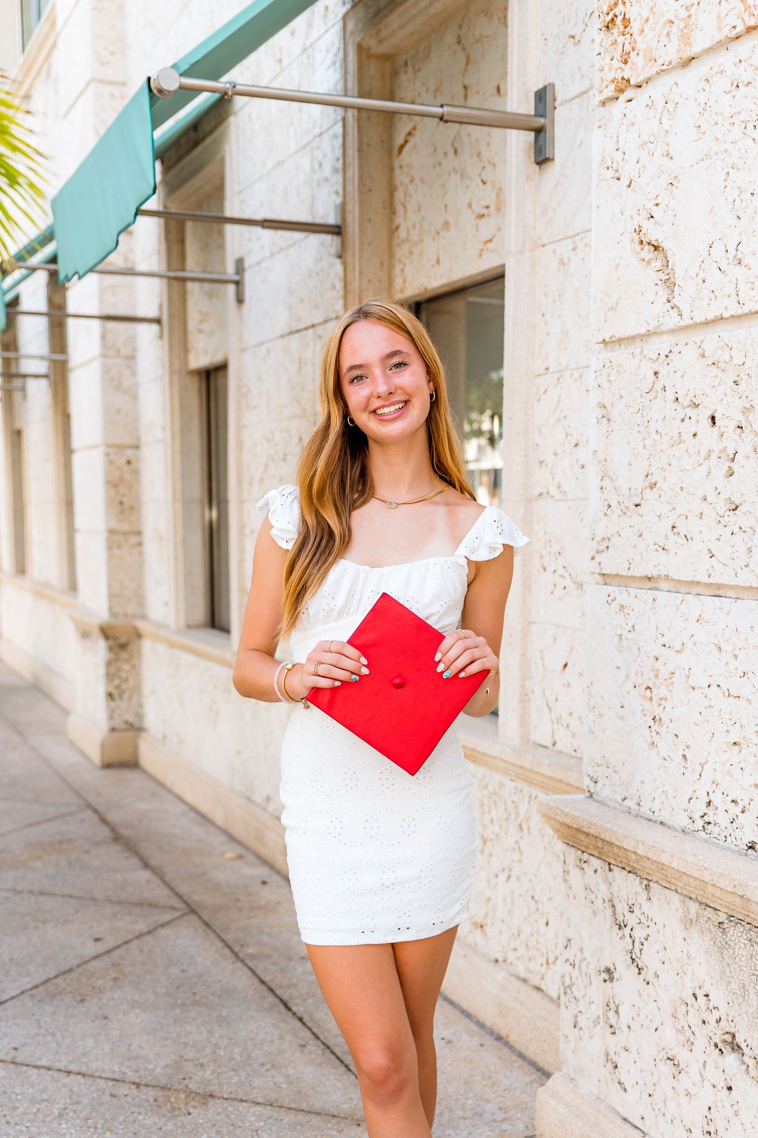 Young woman in a white summer dress holds a red folder while standing outside a stone building.