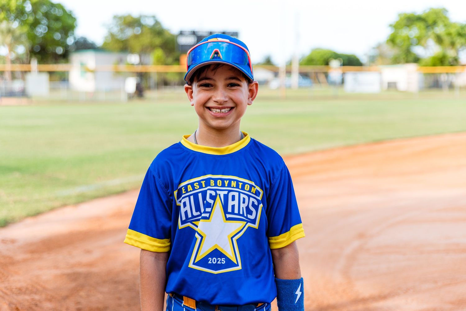 Youth baseball player wearing a bright blue All-Stars jersey and cap smiles on dirt baseball field path.