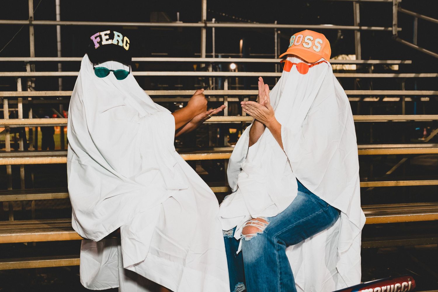 Group in white ghost costumes gather on metal bleachers at night with stadium lights in background.