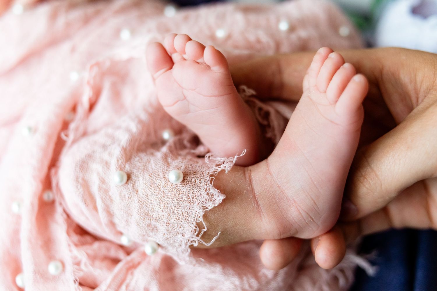 Close-up of a newborn baby's tiny feet held gently in an adult's hand with pink lace fabric.