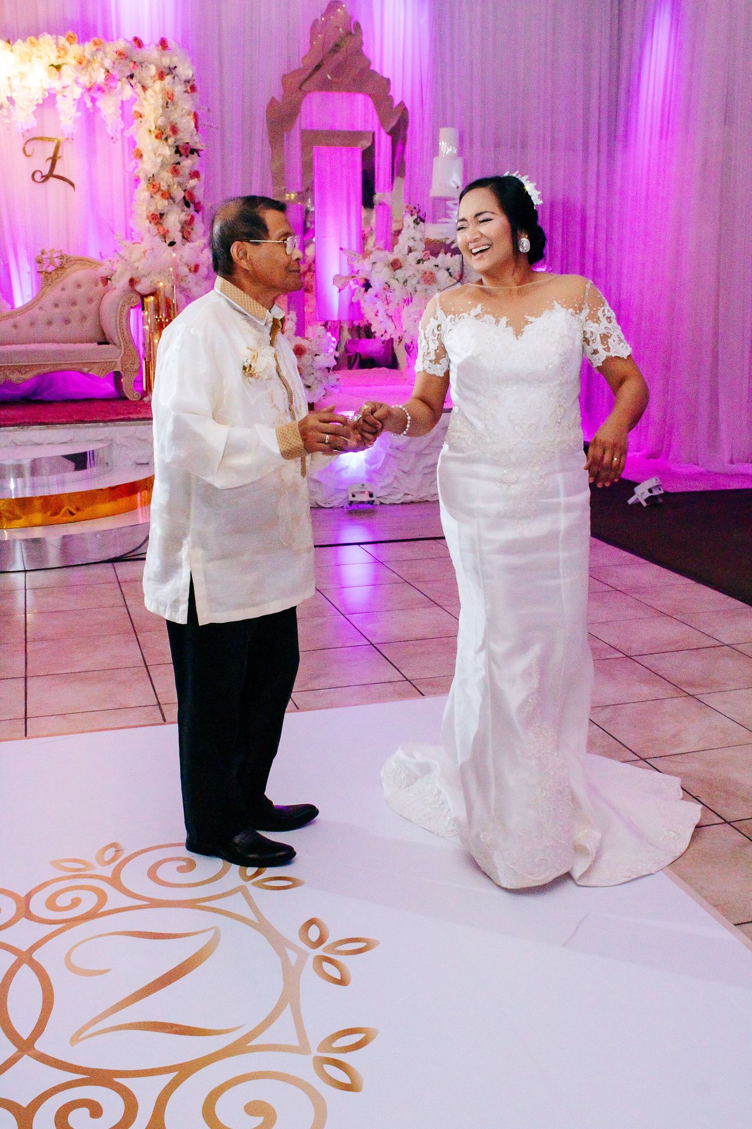 A bride and groom share a romantic dance on an elegant white dance floor with pink lighting and floral decor.