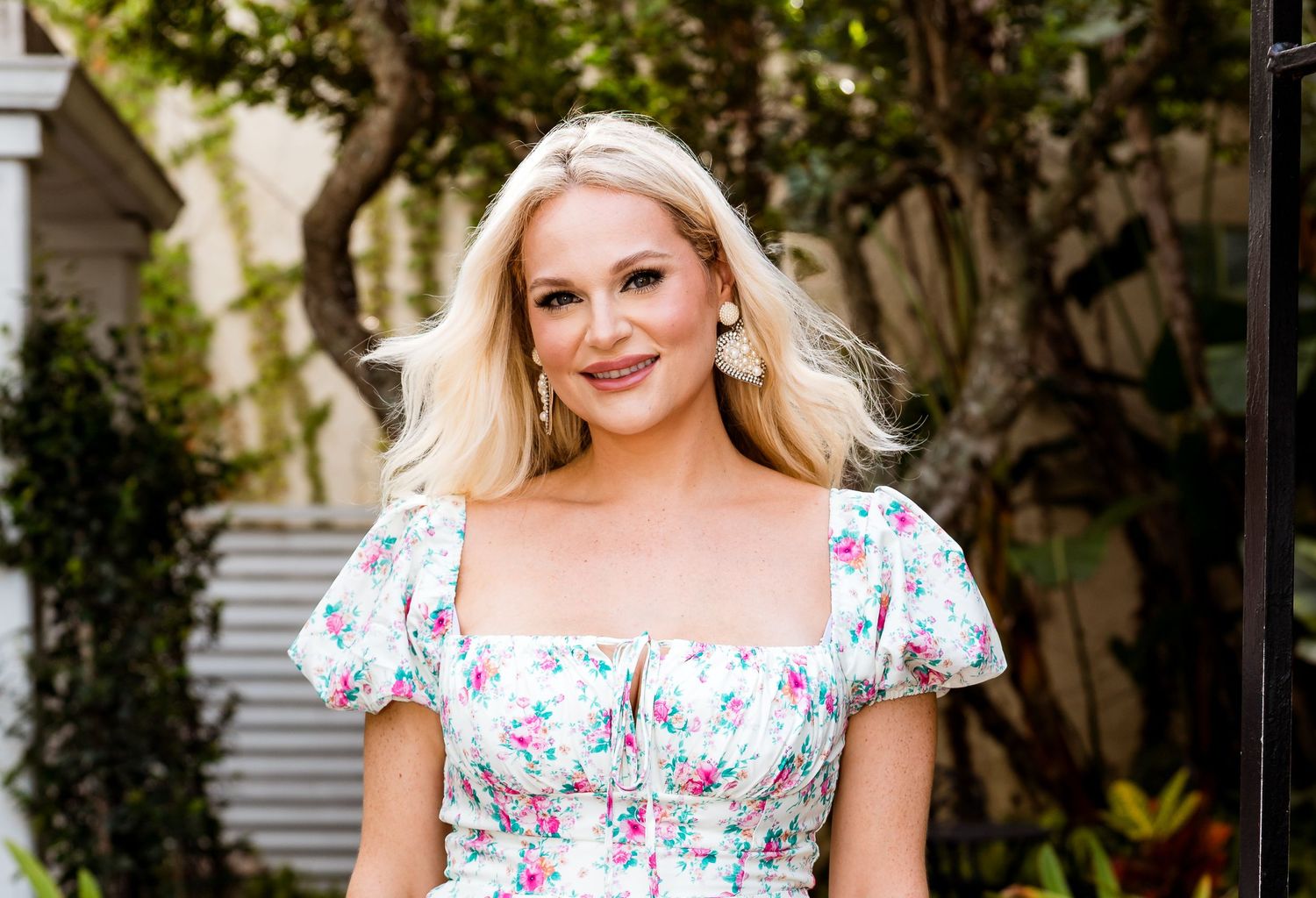 A person in a floral summer dress poses outdoors with greenery in the background.