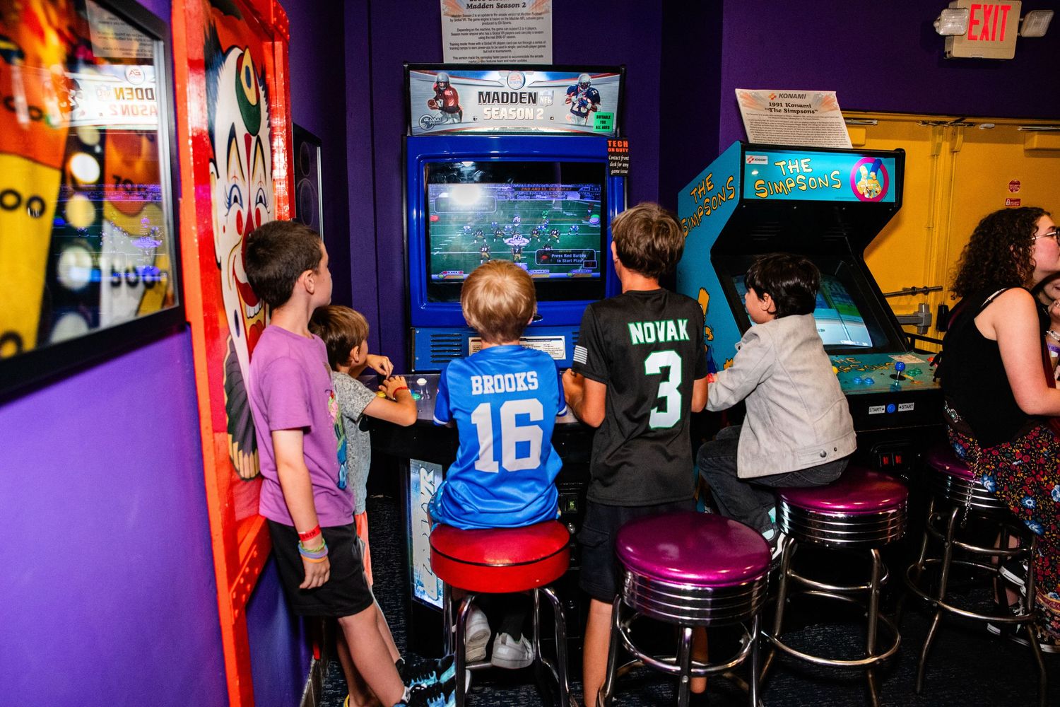 Kids playing arcade video games at a colorful retro arcade bar with purple walls and chrome bar stools.