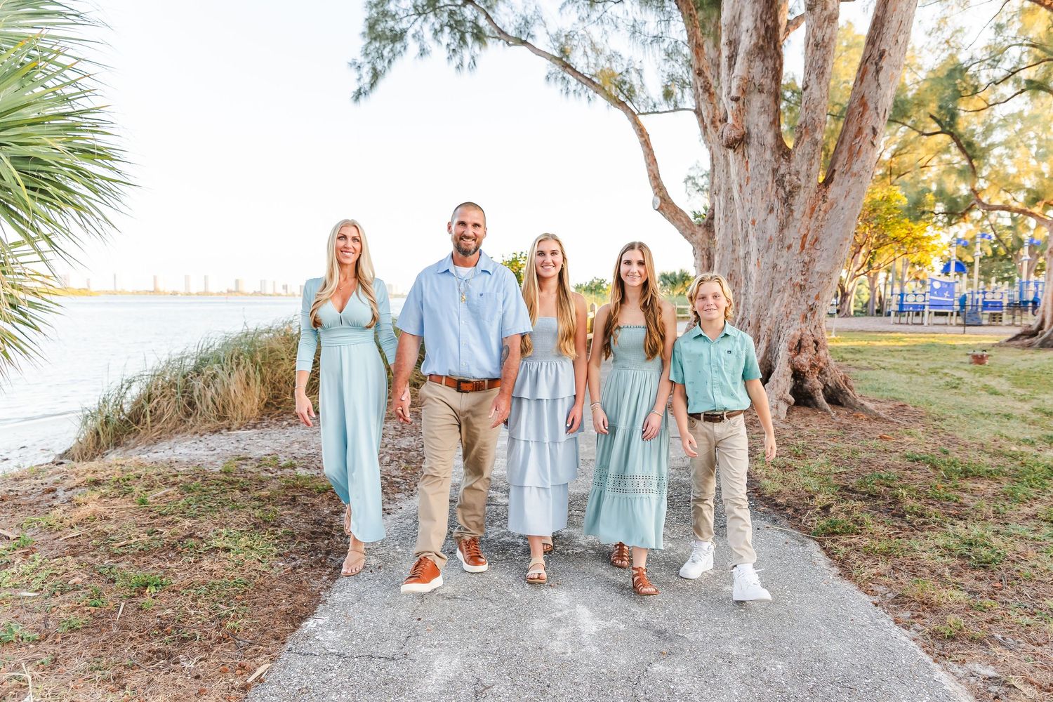 A family wearing coordinated light blue and white outfits poses together on a coastal path with trees and water in background.