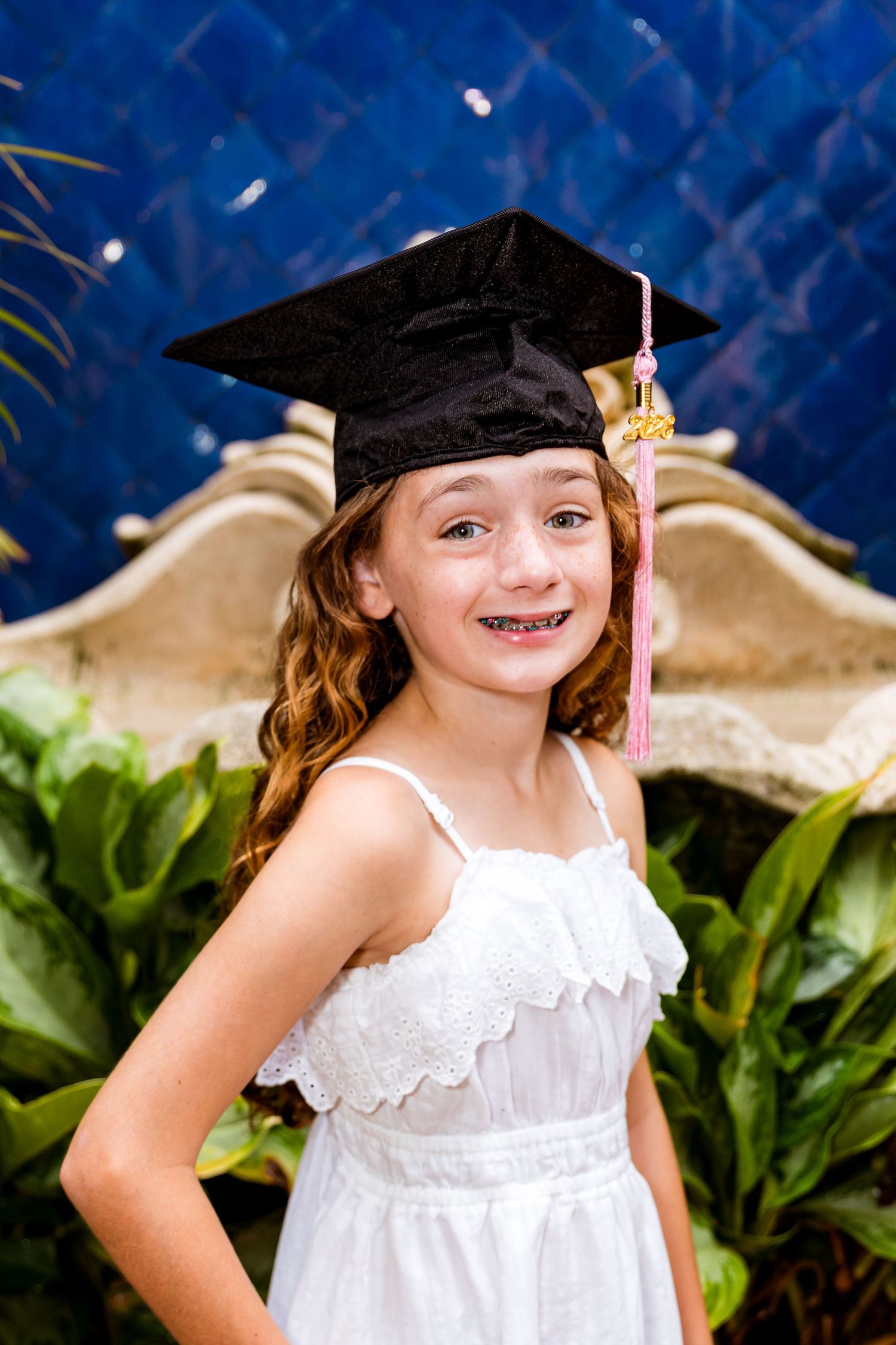 Young girl in white dress and black graduation cap smiles proudly, posing in front of lush green plants.