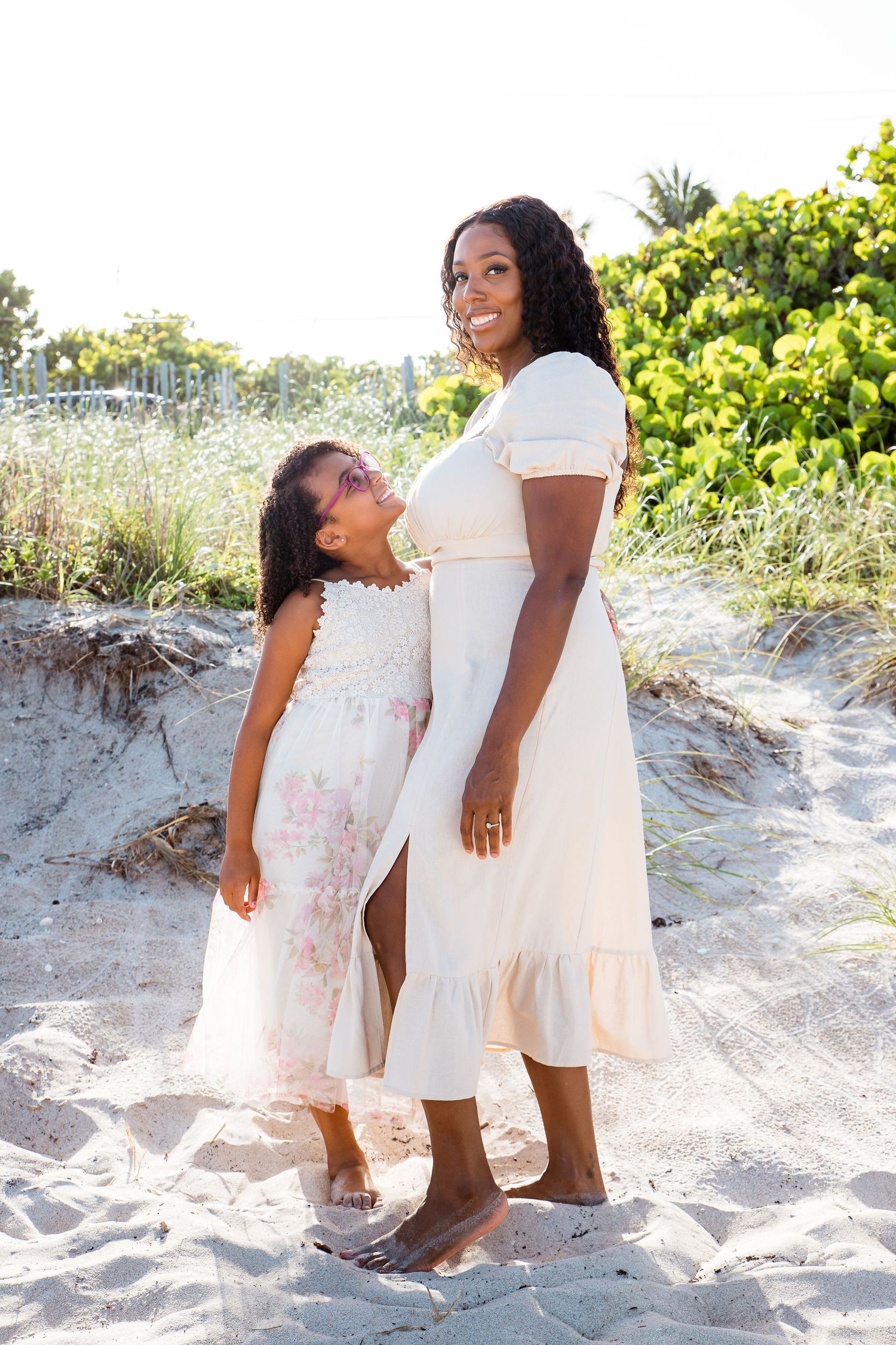 Two figures in light dresses play together at the beach, creating a heartwarming scene on the sandy shore.