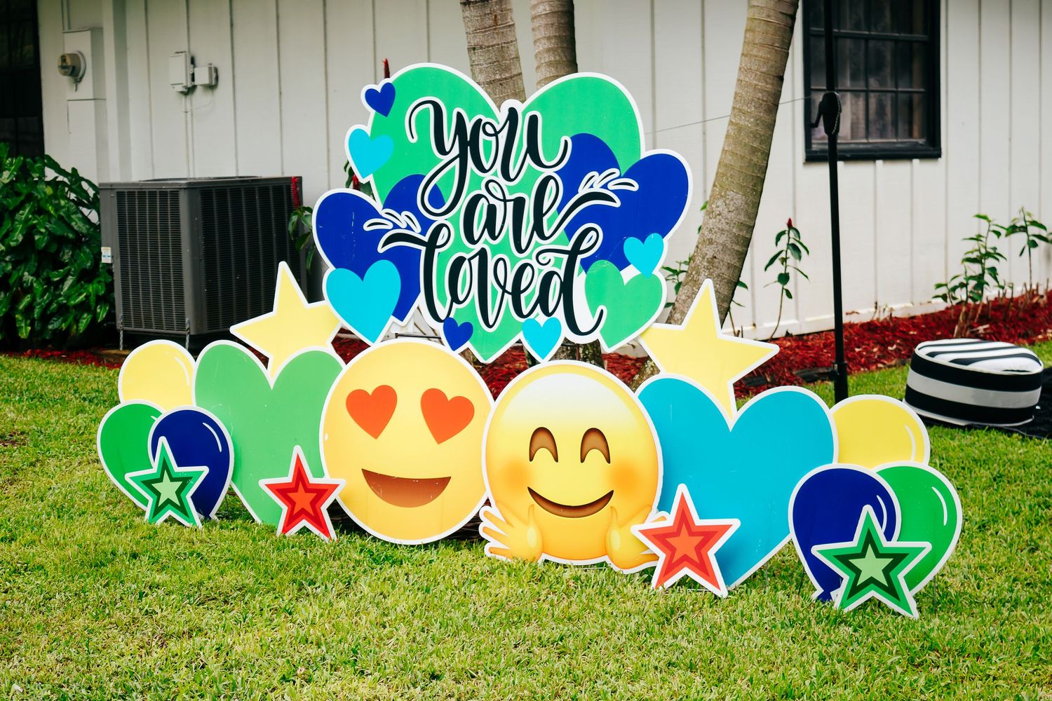 Colorful yard sign display with emoji faces, stars, and 'You are loved' text on green lawn.