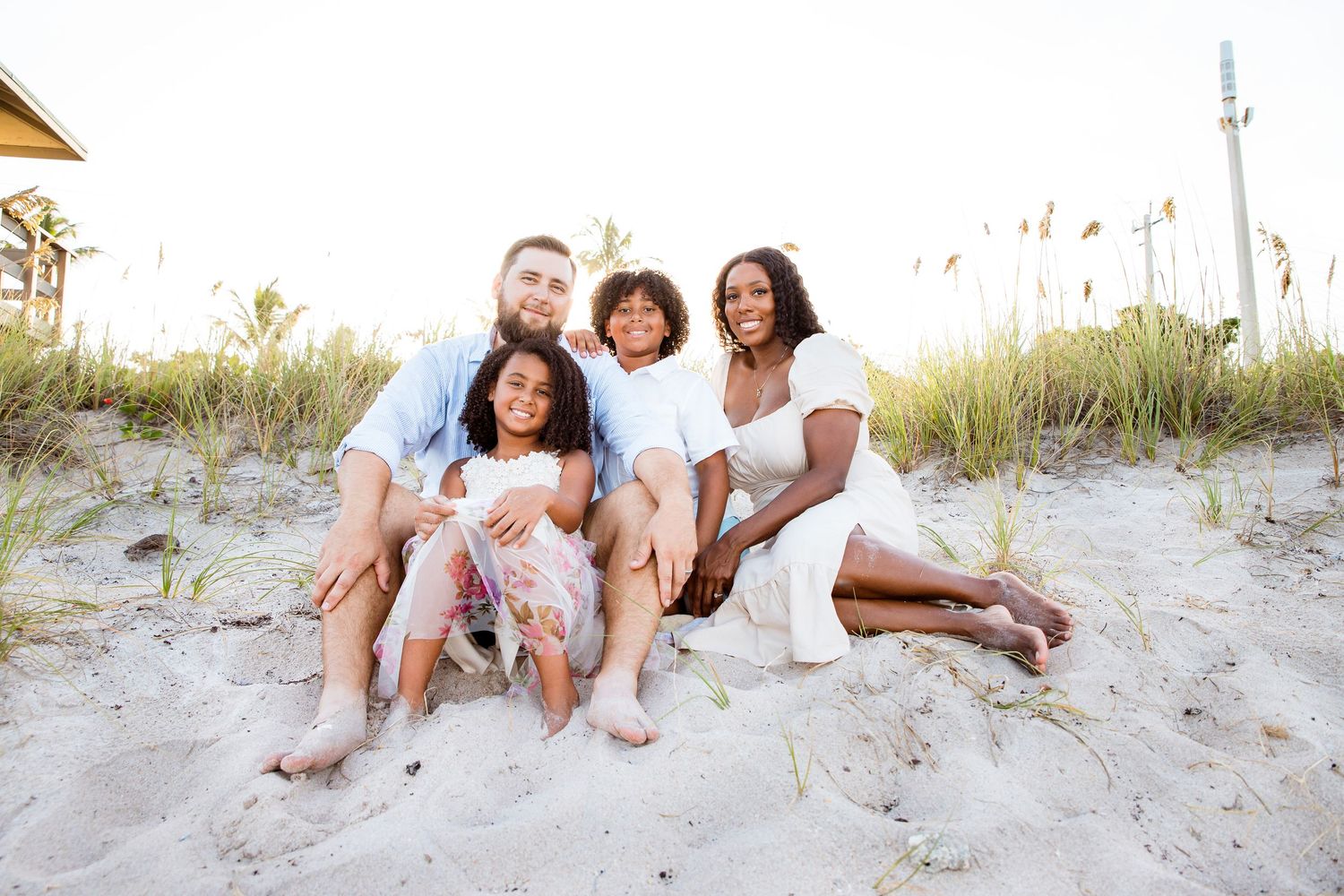 A family sits together smiling on a sandy beach during golden hour with sea grass and dunes in the background.