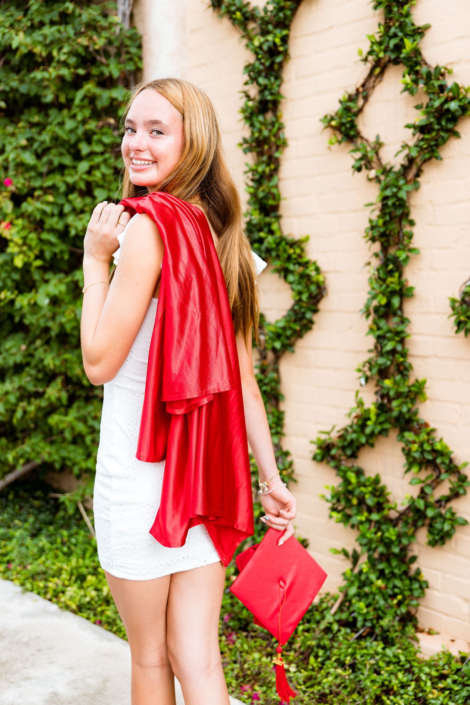 Person in white dress poses against ivy-covered wall while holding red scarf and matching handbag.