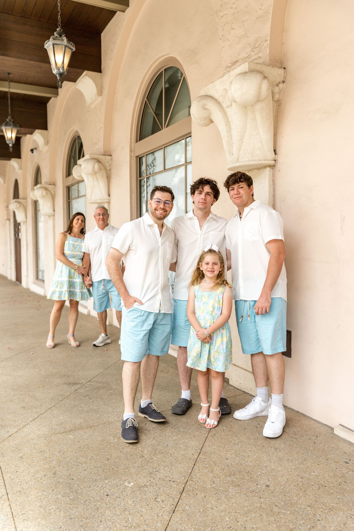 A group in matching white and light blue outfits stands together in a Mediterranean-style colonnade.