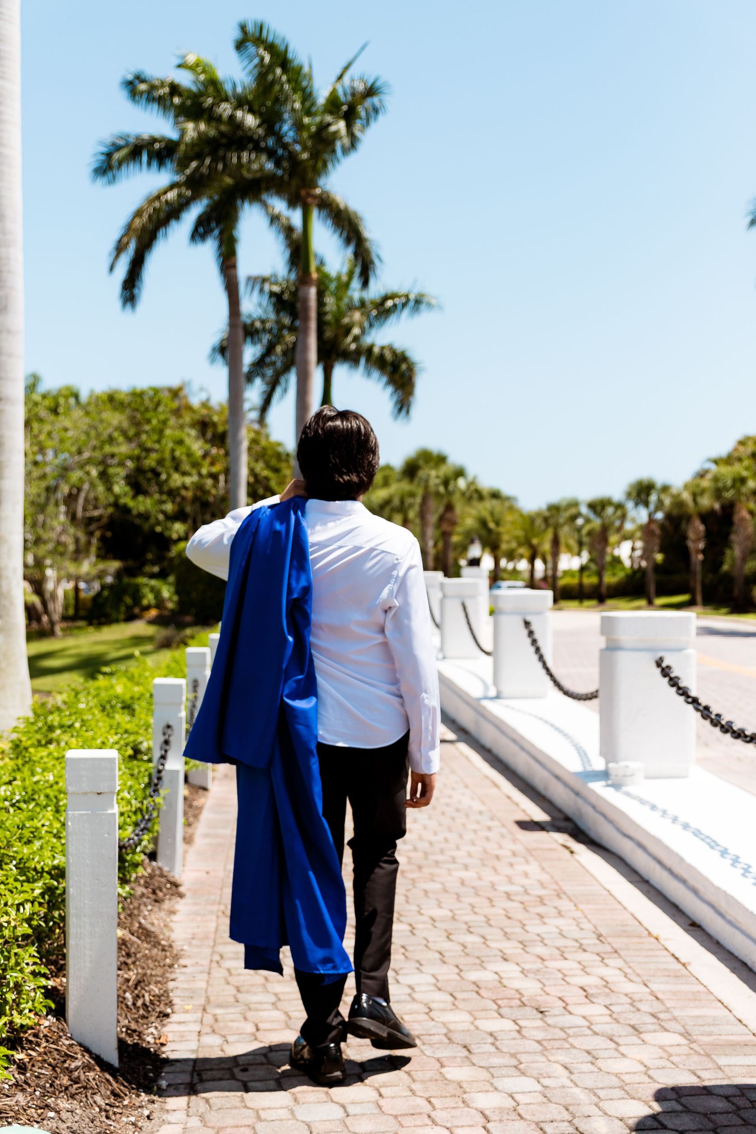 Graduate wearing a blue gown and white shirt walks along a palm-tree lined stone pathway in a tropical setting.