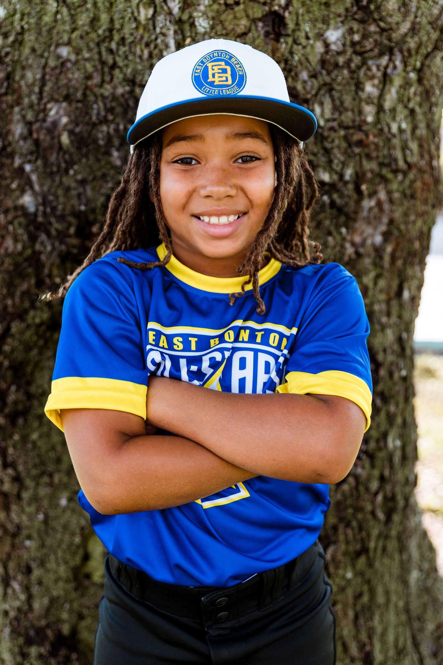 Young athlete wearing baseball uniform in blue and yellow with a white cap poses confidently against tree trunk.
