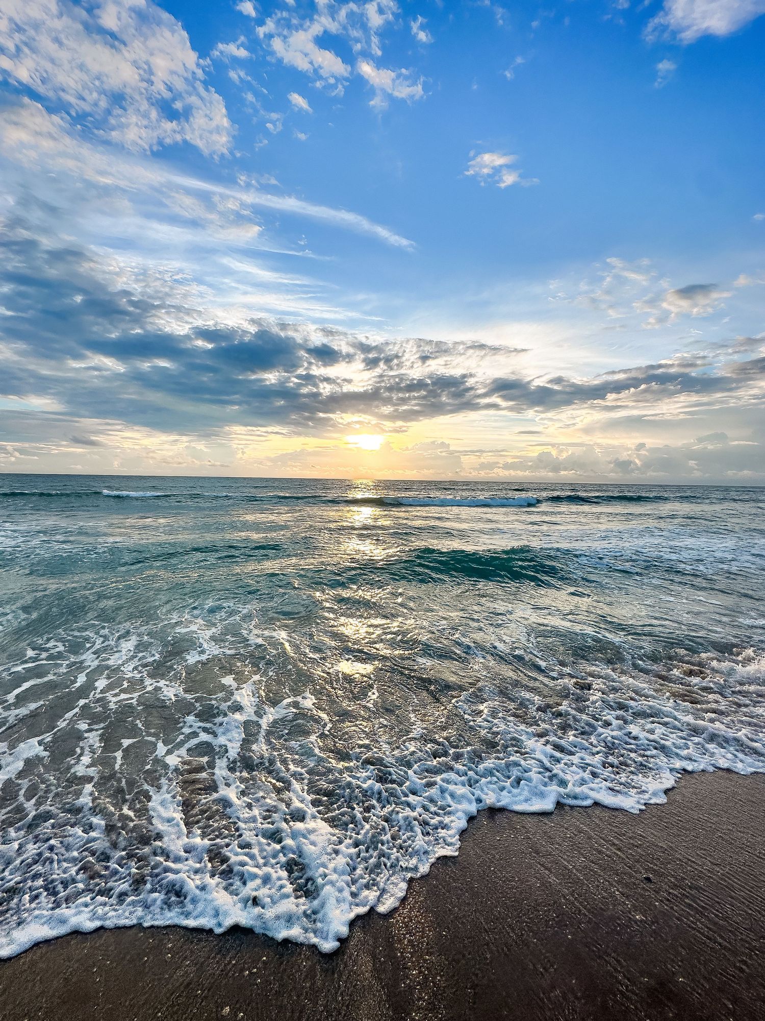 Sunlight pierces through clouds over turquoise ocean waves breaking on sandy beach.