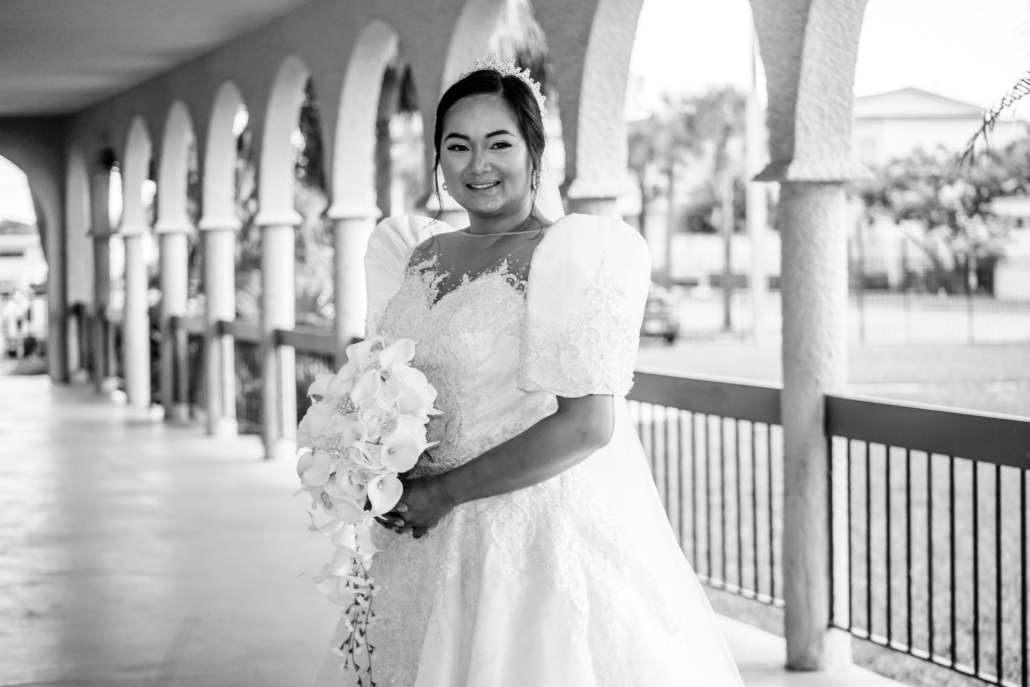 Black and white portrait of bride in elegant dress standing under arched colonnade.