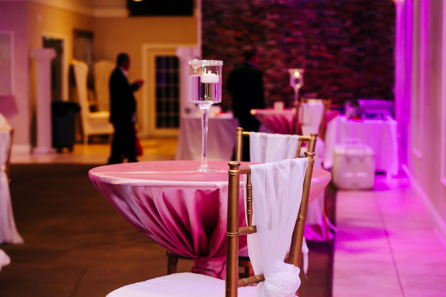 High cocktail tables with white linens and gold decor illuminated by pink uplighting at an elegant event venue.