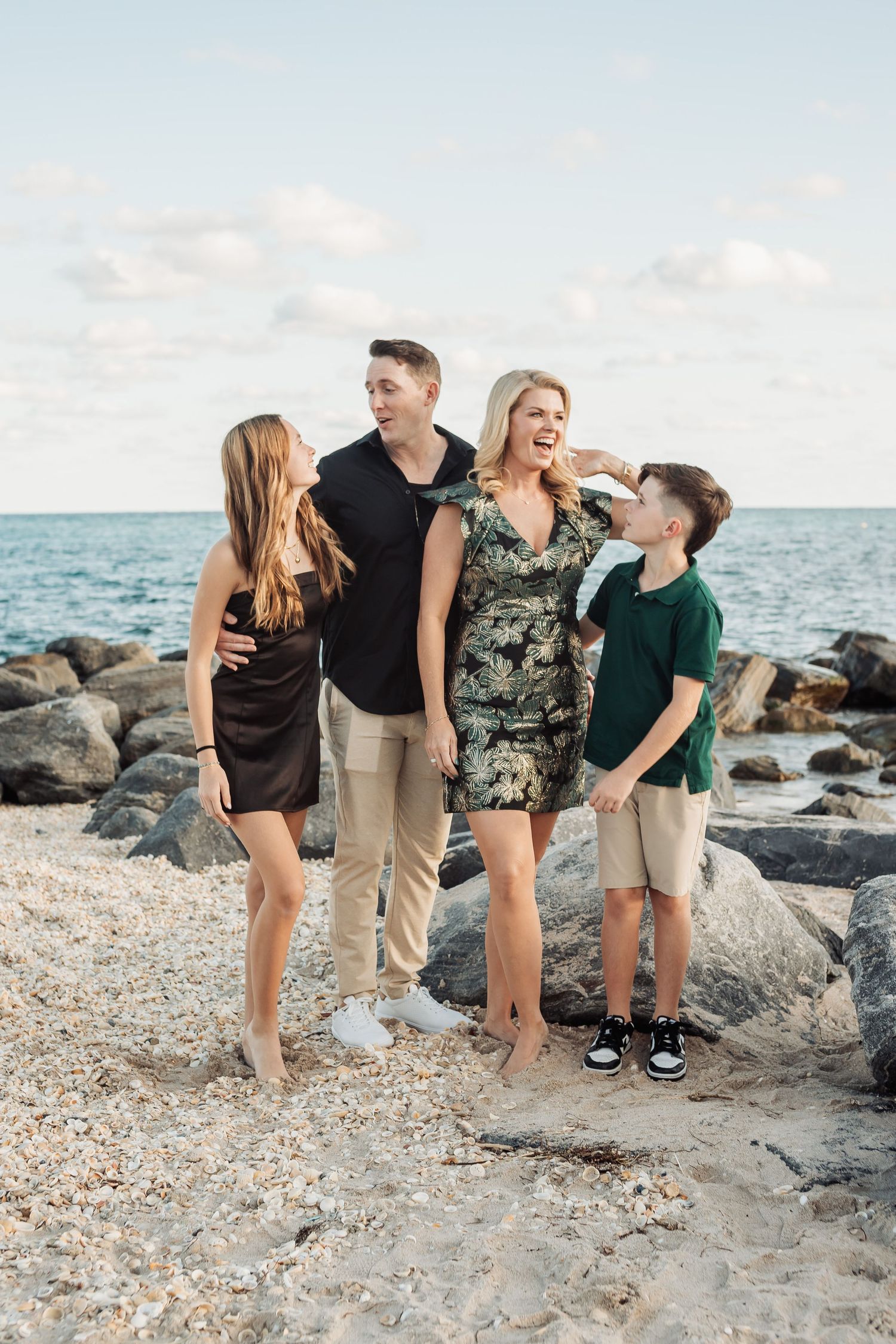 Family shares candid laughing moment together on beach with waves and rocks behind them.
