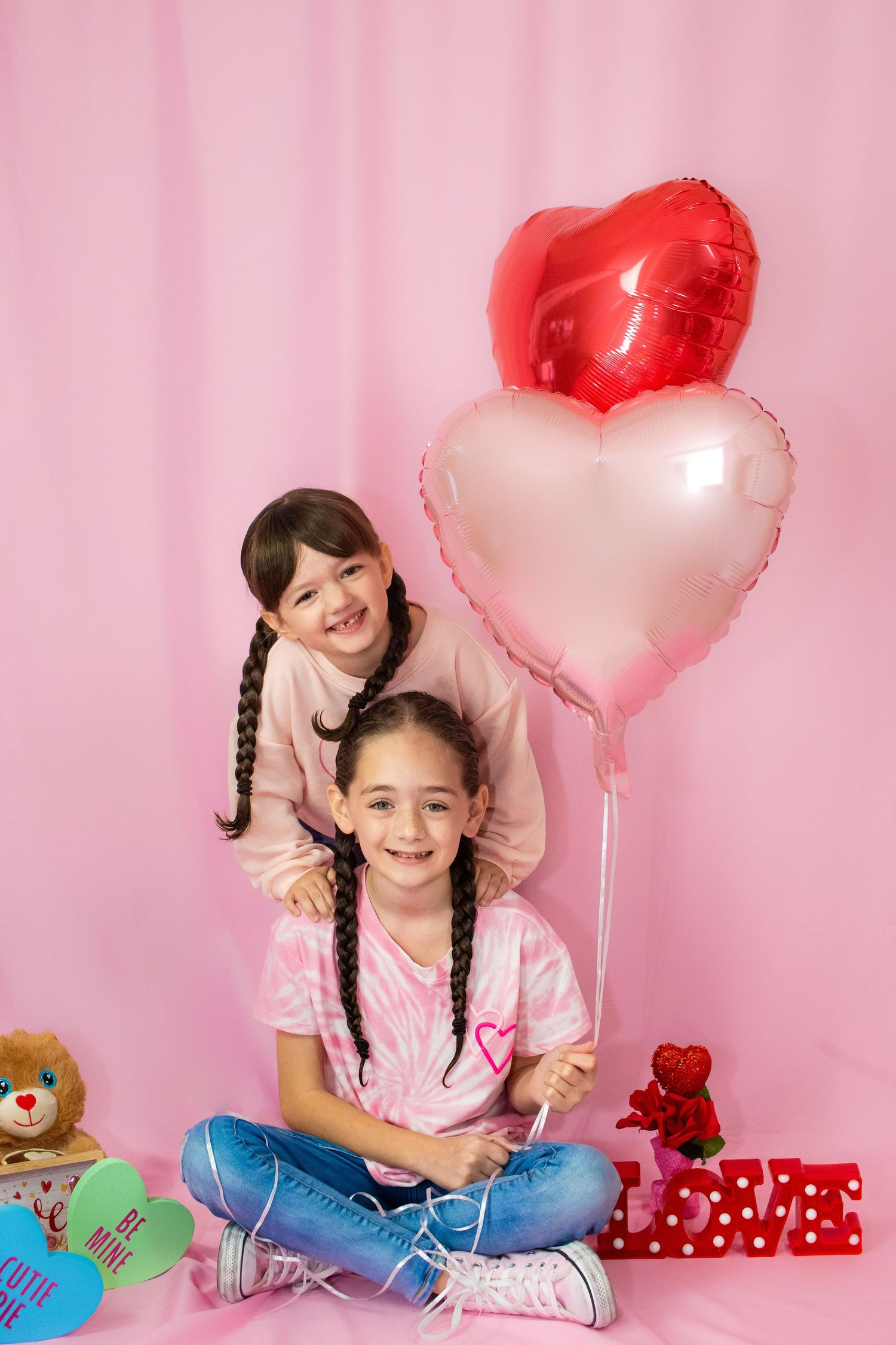 Two children pose playfully with heart-shaped balloons against a pink backdrop with gifts and teddy bears.