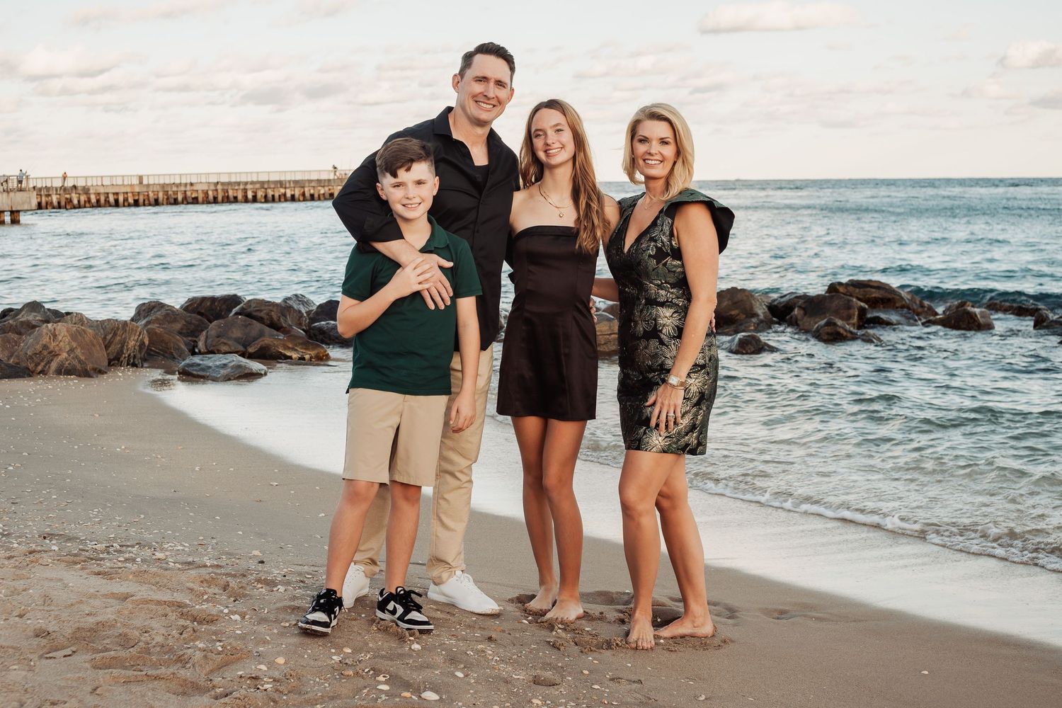 Family poses together on rocky beach with ocean pier in background at sunset.