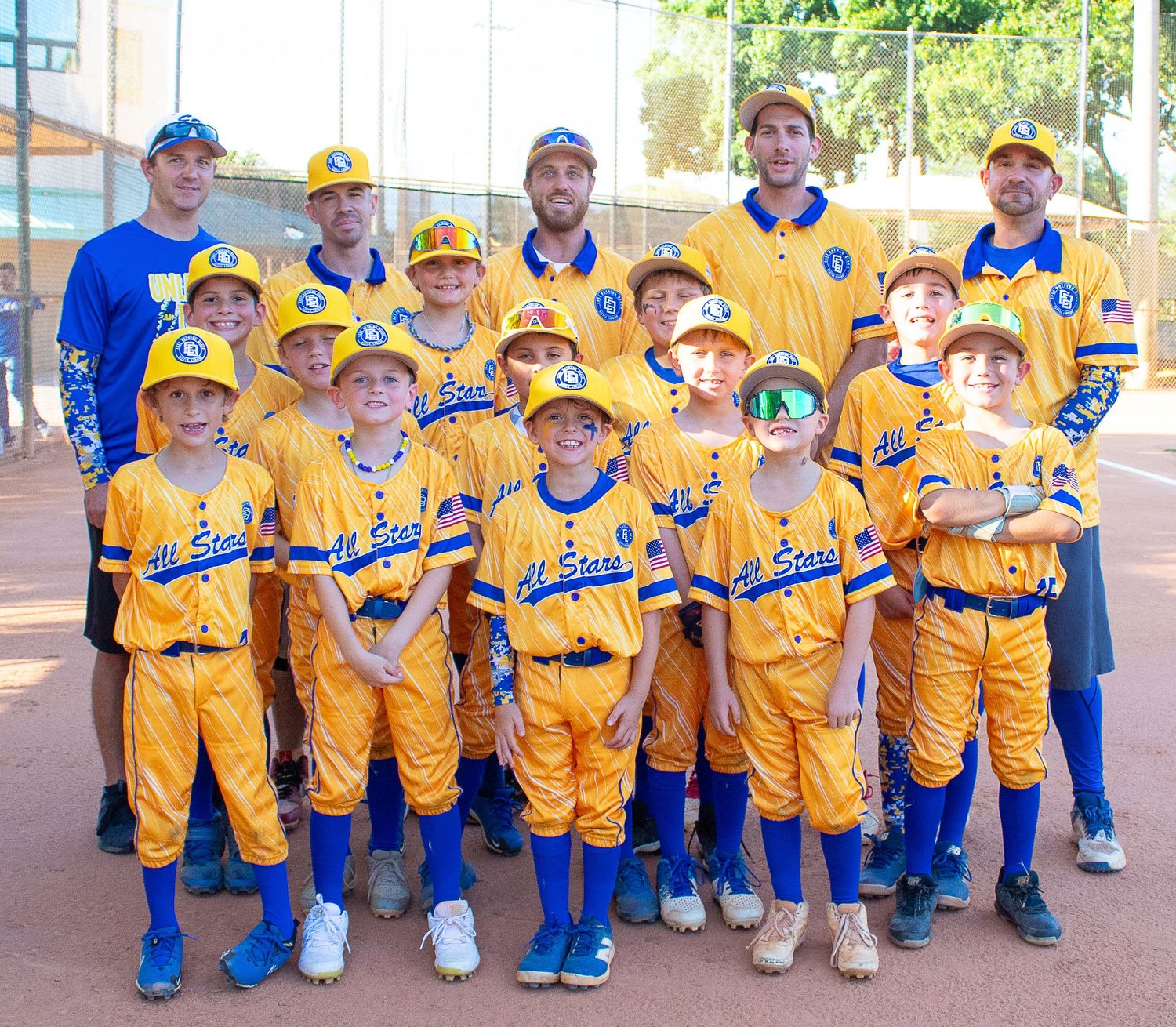 Youth baseball team in bright yellow uniforms and blue accents poses for group photo with coaches on baseball field.