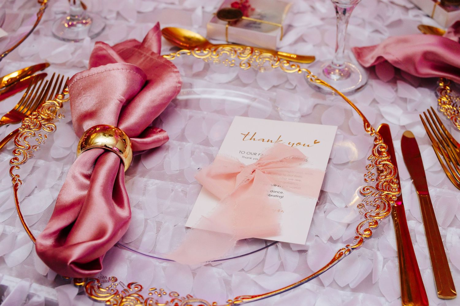 Ornate gold charger plate with pink silk napkin and thank you card on textured tablecloth.