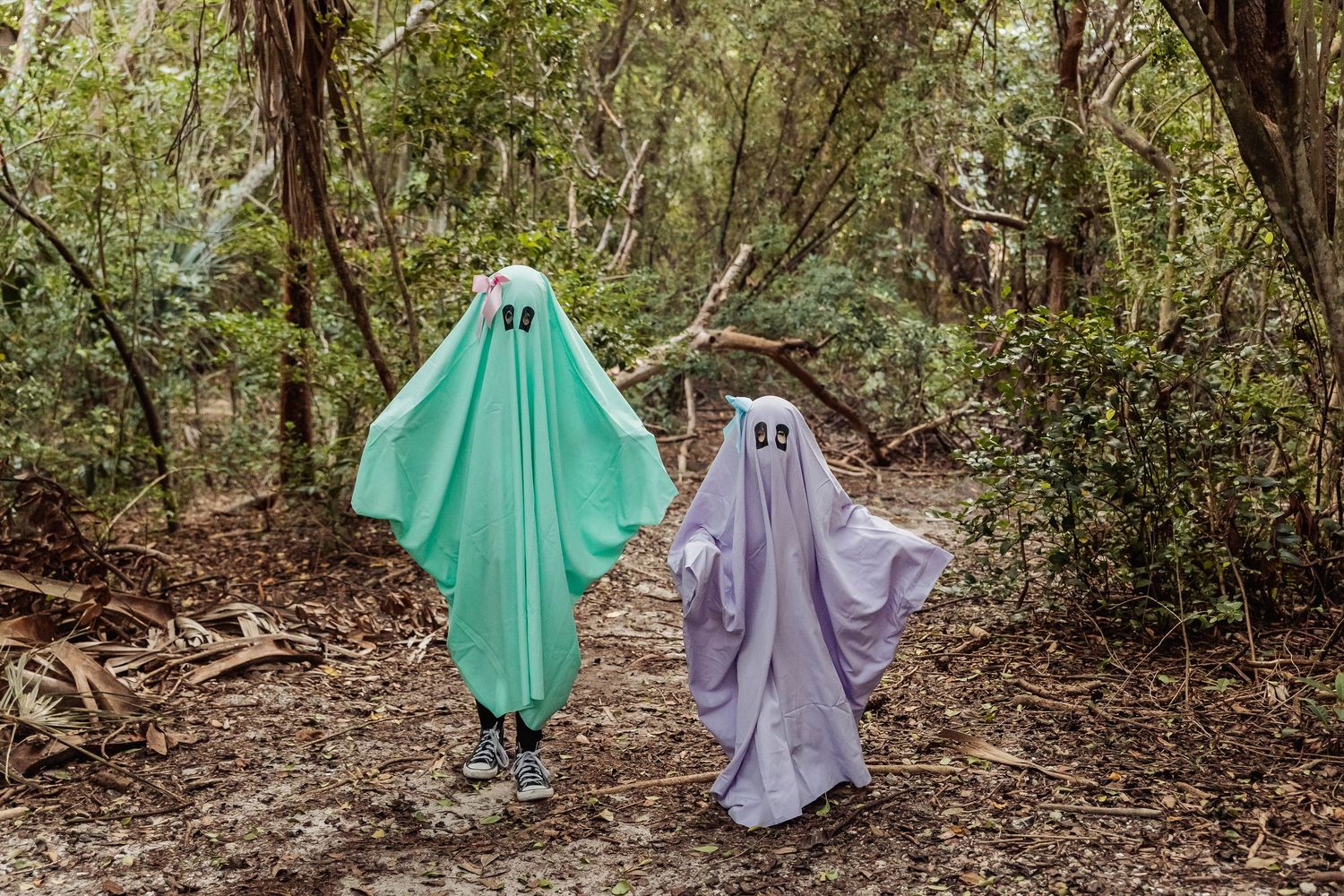Two people in ghost costumes, one green and one purple, pose playfully in a wooded forest setting.