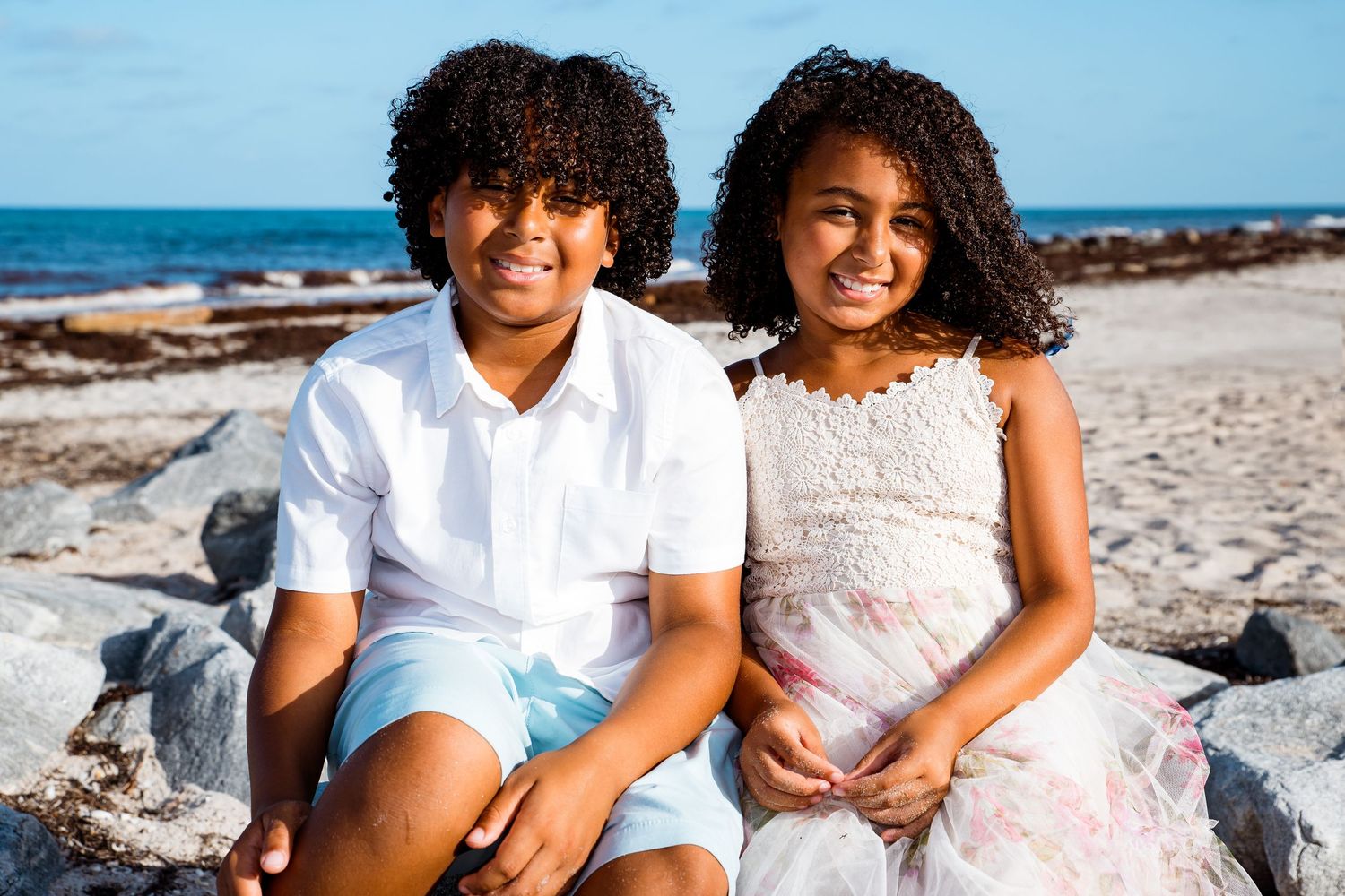 Two children with curly hair sit together smiling on a rocky beach with ocean waves and blue sky in the background.