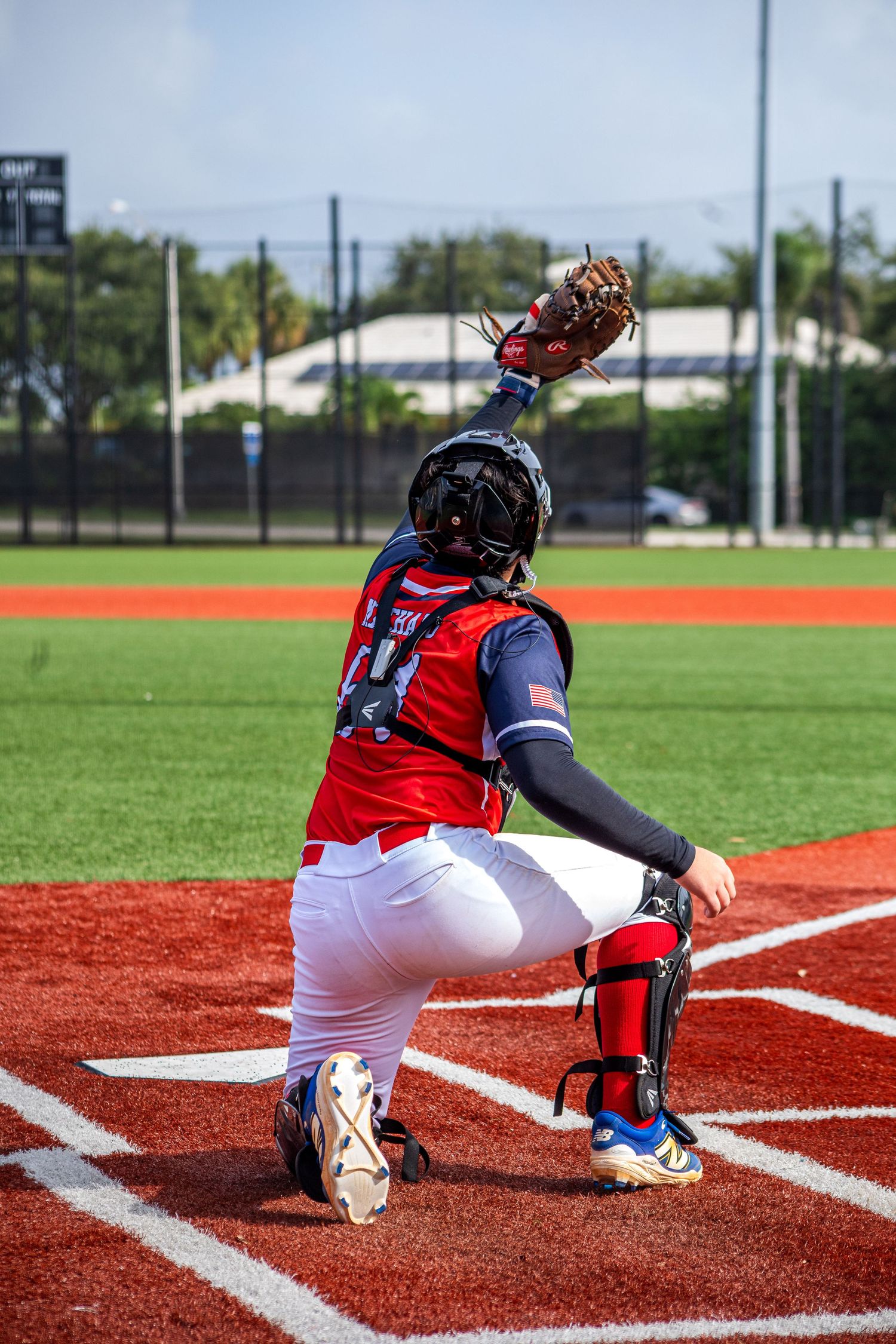 Baseball catcher in red and white uniform crouching at home plate, reaching up with mitt to catch a pitch.
