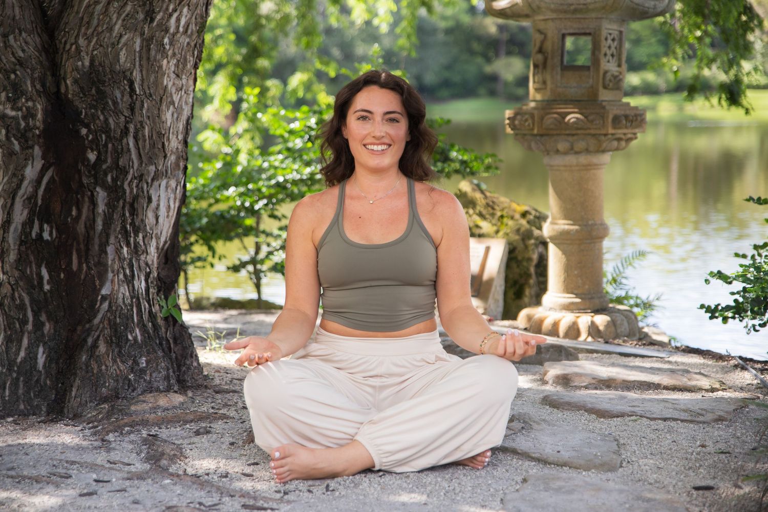 Person in gray athletic top and light pants practicing yoga outdoors by a tranquil pond with stone lantern in background.