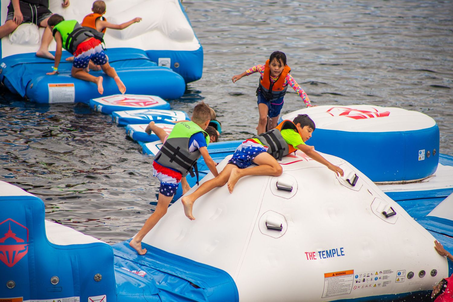 People enjoying a summer day at an aqua park with inflatable blue and white floating platforms on the water.
