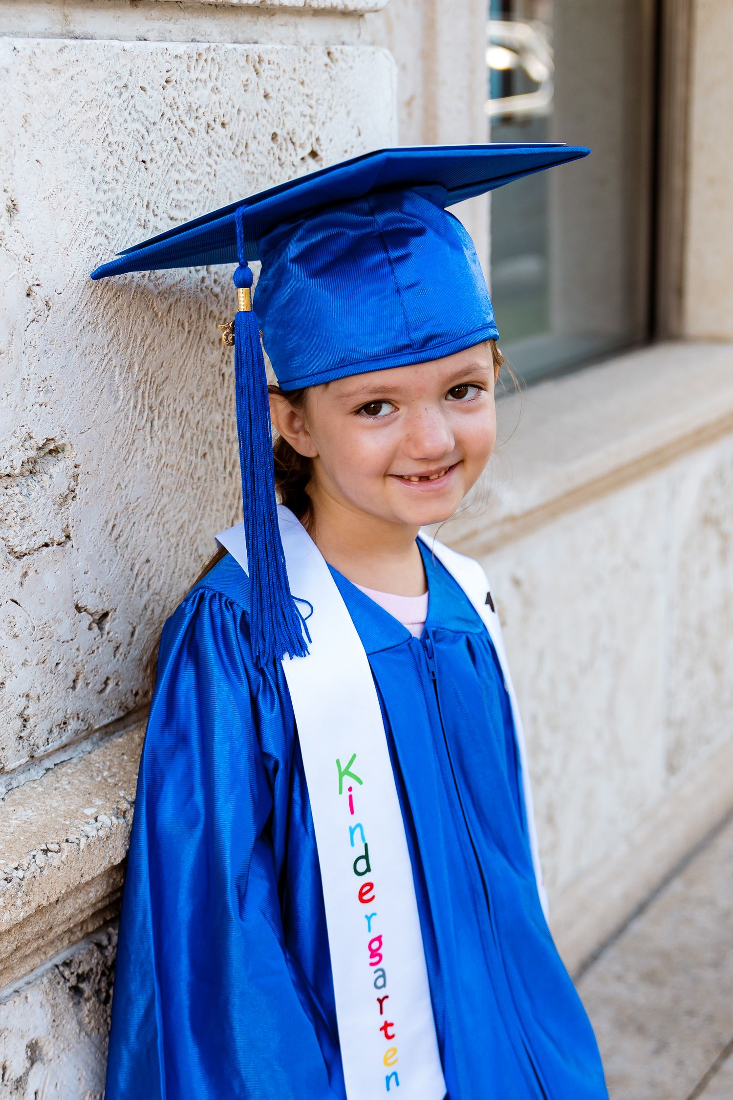 A kindergarten graduate wearing a bright blue cap and gown with a white sash stands against a light colored wall.