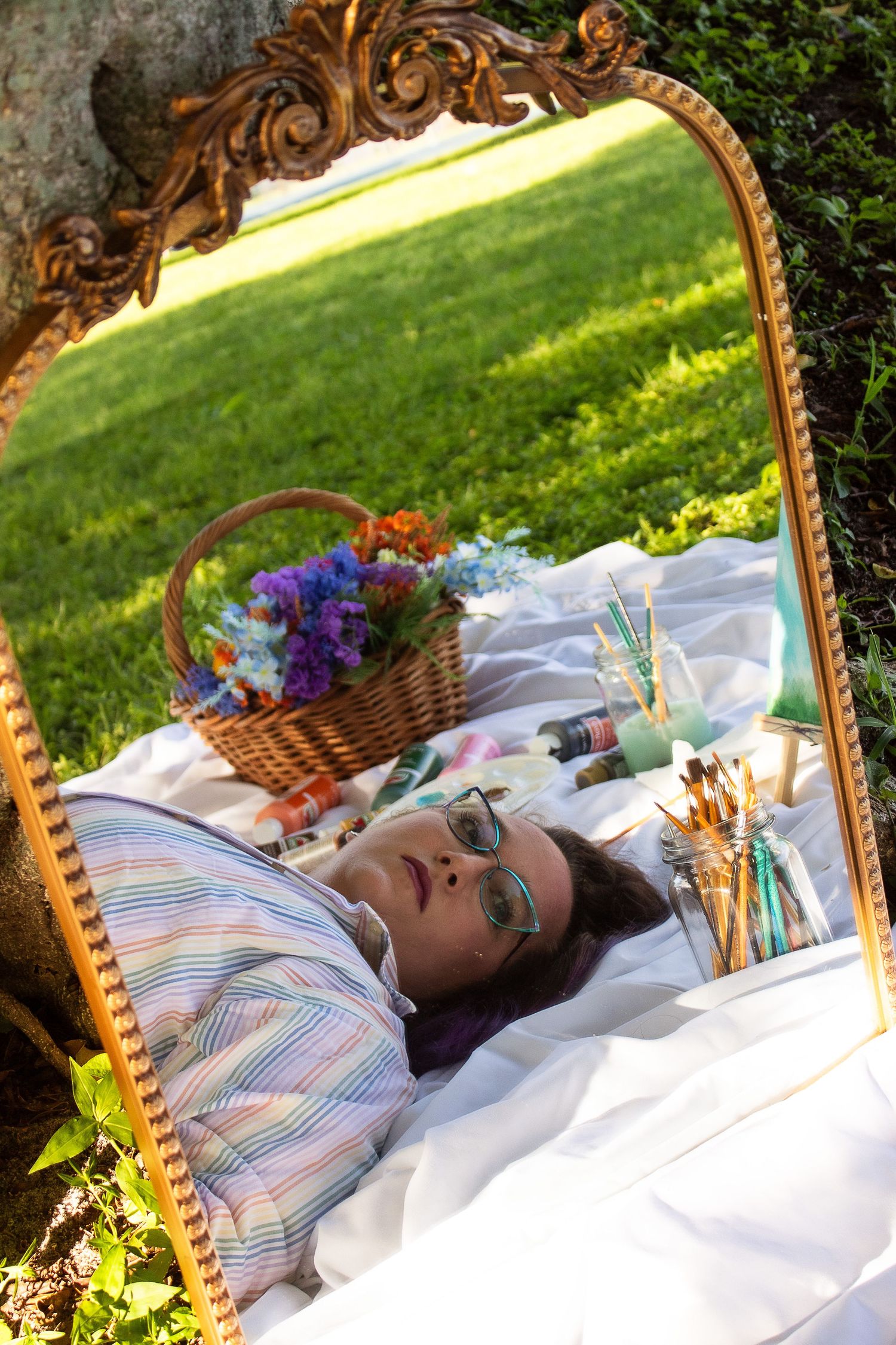 Art supplies and floral basket reflected in vintage gold mirror on white blanket in sunny garden.
