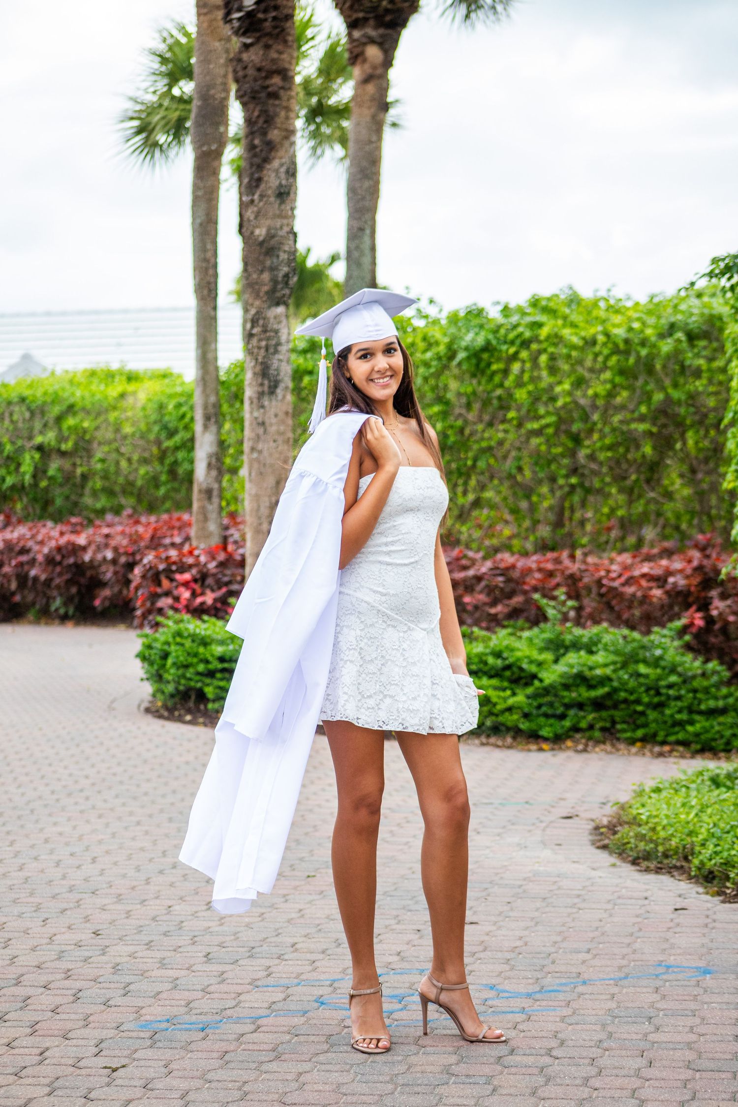 A woman in a white dress and hat stands in a tropical garden setting with palm trees and lush greenery.